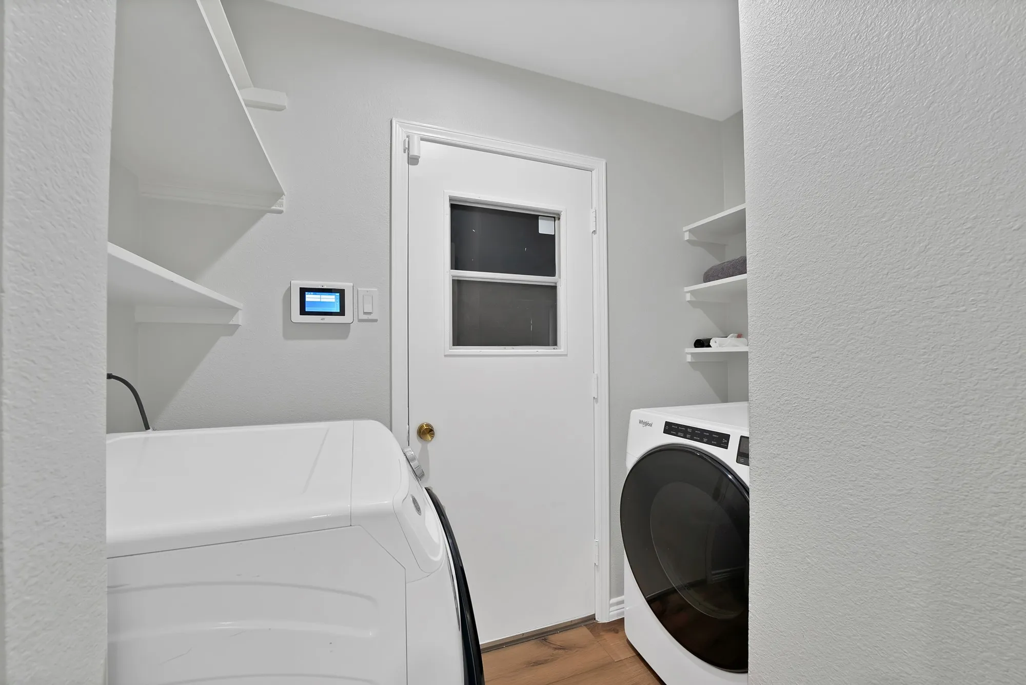 Dedicated laundry room featuring front-load washer and dryer, white shaker-style cabinetry with open shelving, wood-finish flooring, and a white paneled exit door with transom window for natural light and ventilation