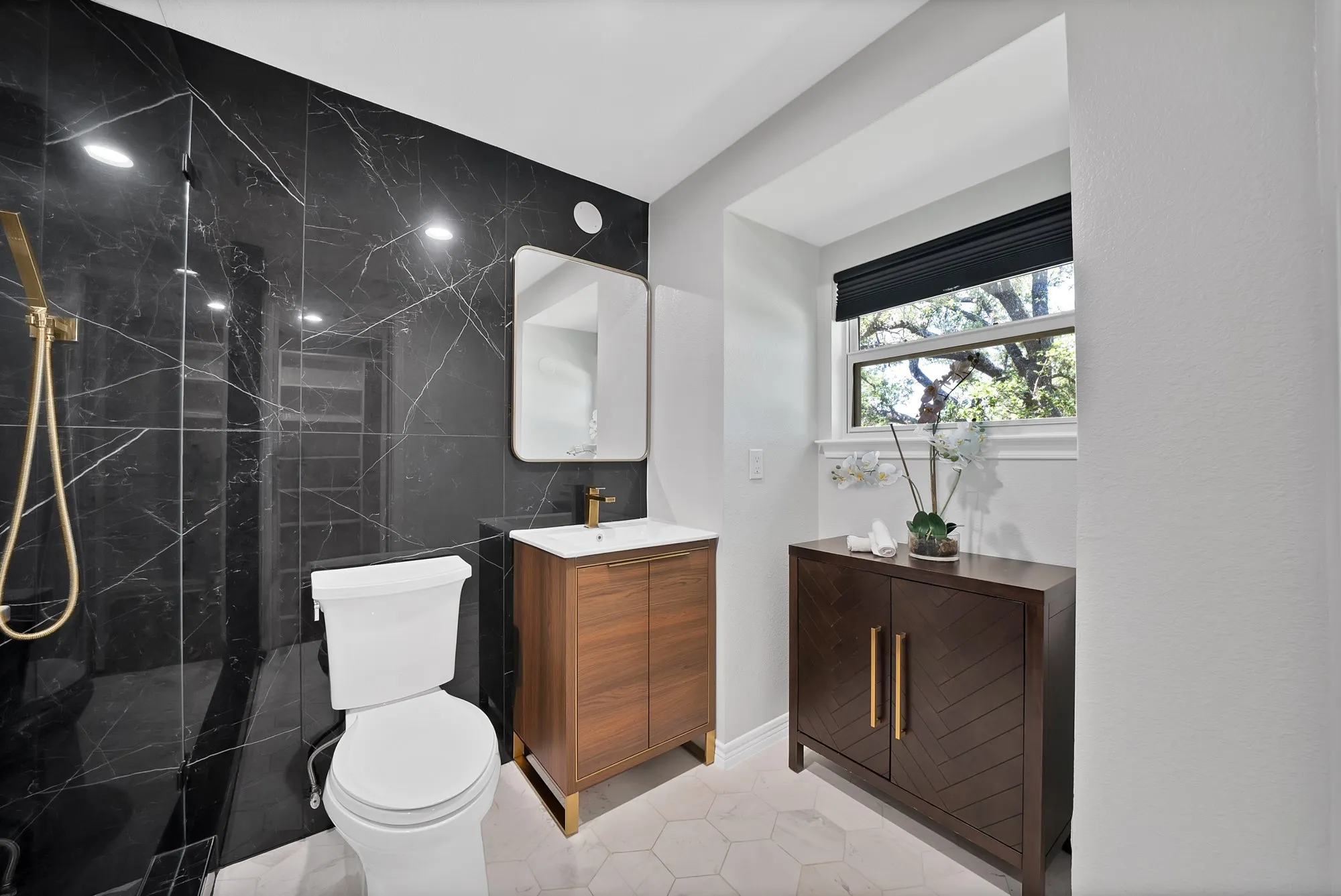 Contemporary primary bathroom featuring a walk-in shower with black marble-look tile surround and glass enclosure, floating wood-finish vanity with integrated sink and brass hardware, round mirror with integrated lighting, and light marble-look flooring