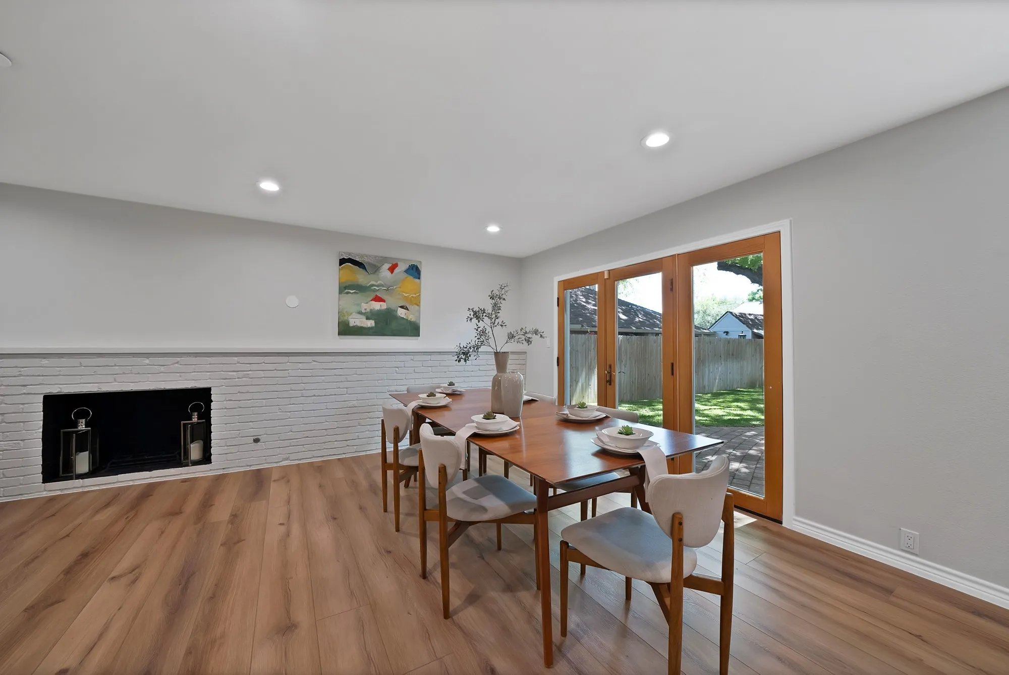 Wood-finish flooring, recessed lighting, white painted brick fireplace, and wood-framed French doors opening to a paved patio and fenced yard