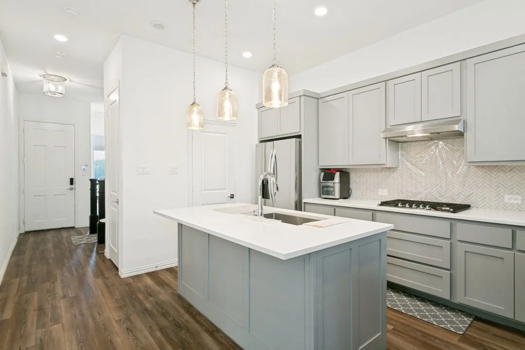 Contemporary kitchen featuring soft-gray cabinetry with brushed stainless steel appliances, white quartz countertops, a waterfall-edge island, pendant lighting, herringbone tile backsplash, and wood-finish flooring throughout the open layout