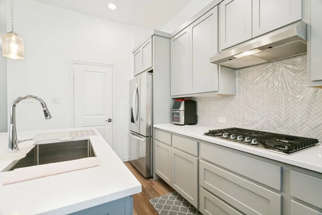 Modern kitchen featuring light gray cabinetry with integrated hardware, white quartz countertops, stainless steel appliances including a gas cooktop and under-cabinet range hood, herringbone subway tile backsplash, and a waterfall-edge island with undermount sink and pull-down faucet