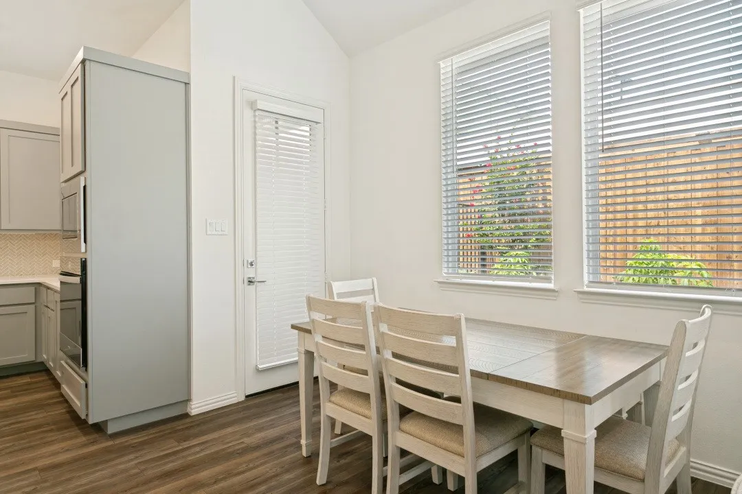 Bright kitchen and dining area featuring wood-finish flooring, horizontal blinds across expansive windows, white cabinetry with integrated appliances, and a light wood dining table with seating for four