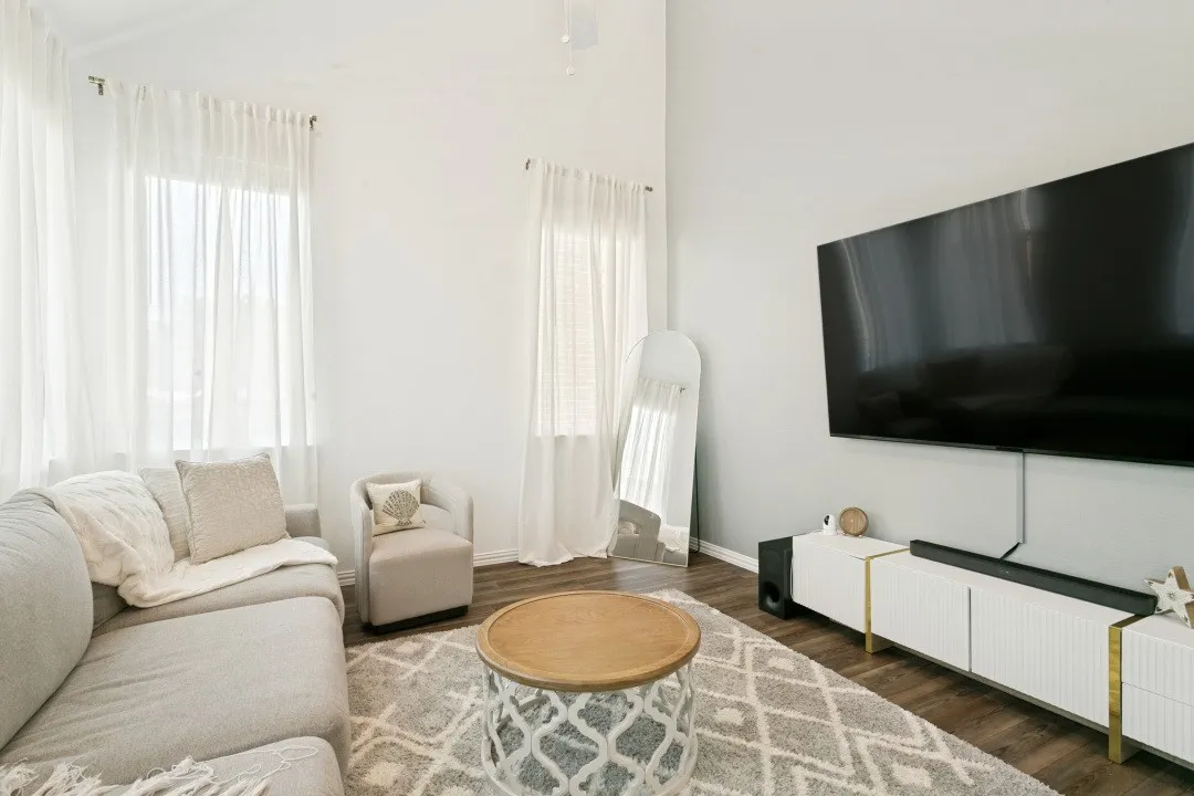 Bright living room featuring floor-to-ceiling windows with sheer curtains, hardwood flooring, a round wood-top accent table, geometric area rug, wall-mounted flat-screen television, and white cabinetry with brass hardware