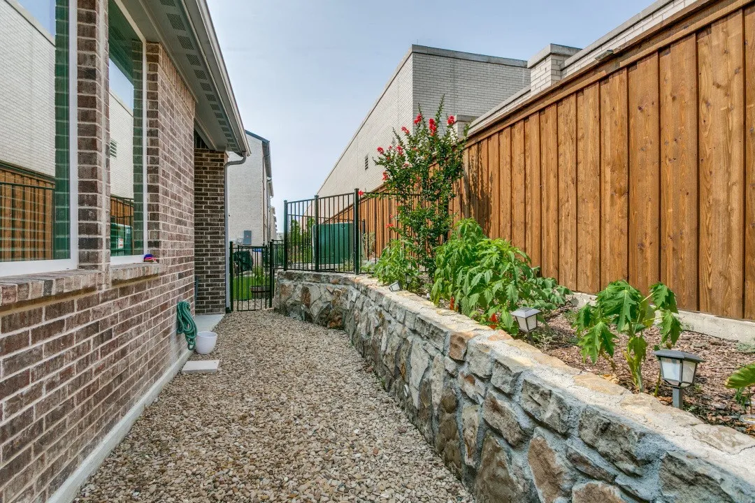 Brick exterior with white-framed windows, gravel walkway, stone retaining wall, and wood privacy fence with a black metal gate
