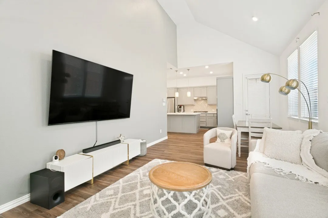 Open-concept living space featuring a wall-mounted flat-screen television, low-profile media console with storage, wood-finish flooring throughout, and abundant natural light from multiple windows