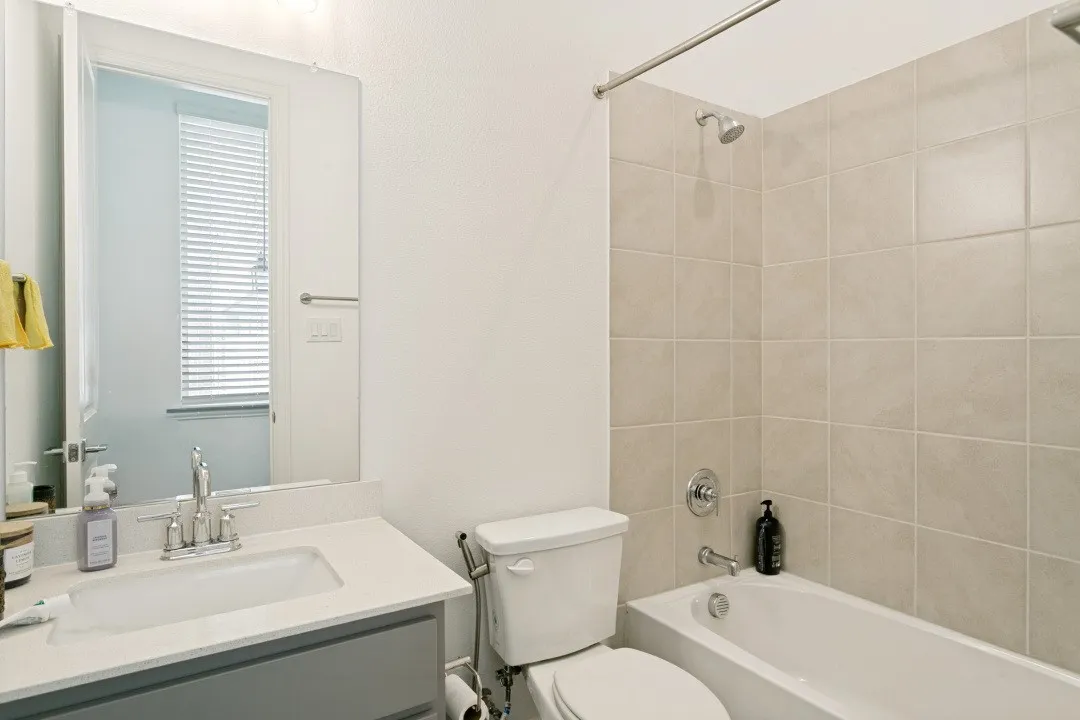 Clean and functional bathroom featuring a gray vanity with white countertop and integrated sink, ceramic tile surround in the shower/tub area, chrome fixtures, and a large mirror with natural light from the window
