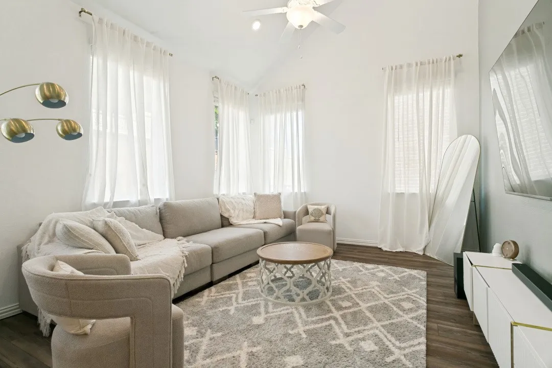 Bright, light-filled living room featuring expansive windows with sheer curtains, wood-finish flooring, and a geometric patterned area rug