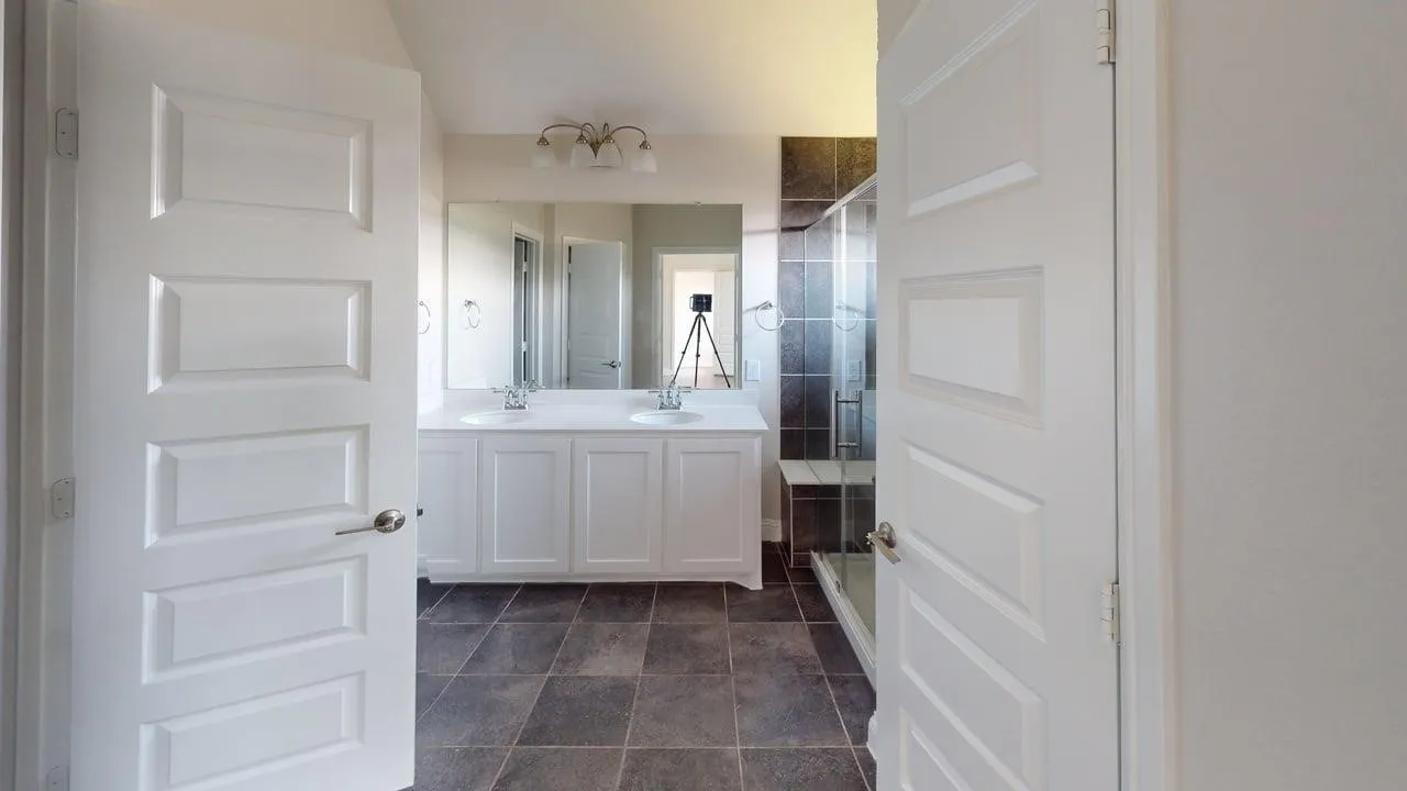 White paneled doors opening to a bathroom featuring a double vanity with dual sinks, tiled flooring, and a glass-enclosed shower with tiled walls and a built-in bench