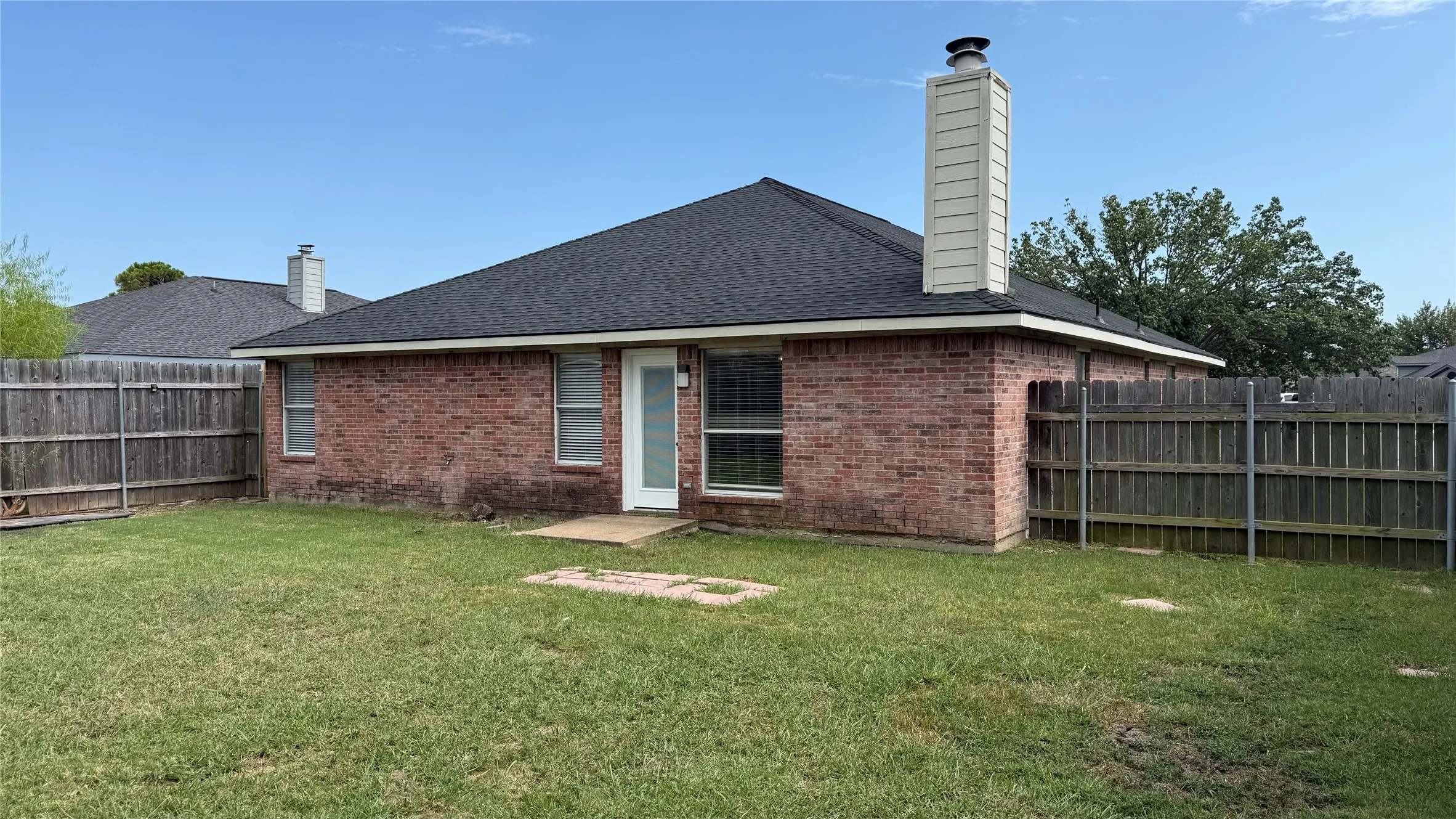 Brick ranch-style home featuring a gabled roof with tall chimney, rear patio area, and expansive fenced yard with mature landscaping