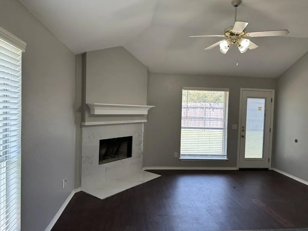 Spacious living area with wood-finish flooring, a focal-point fireplace with stone surround and built-in shelving, ceiling fan with integrated lighting, and glass door access to outdoor space