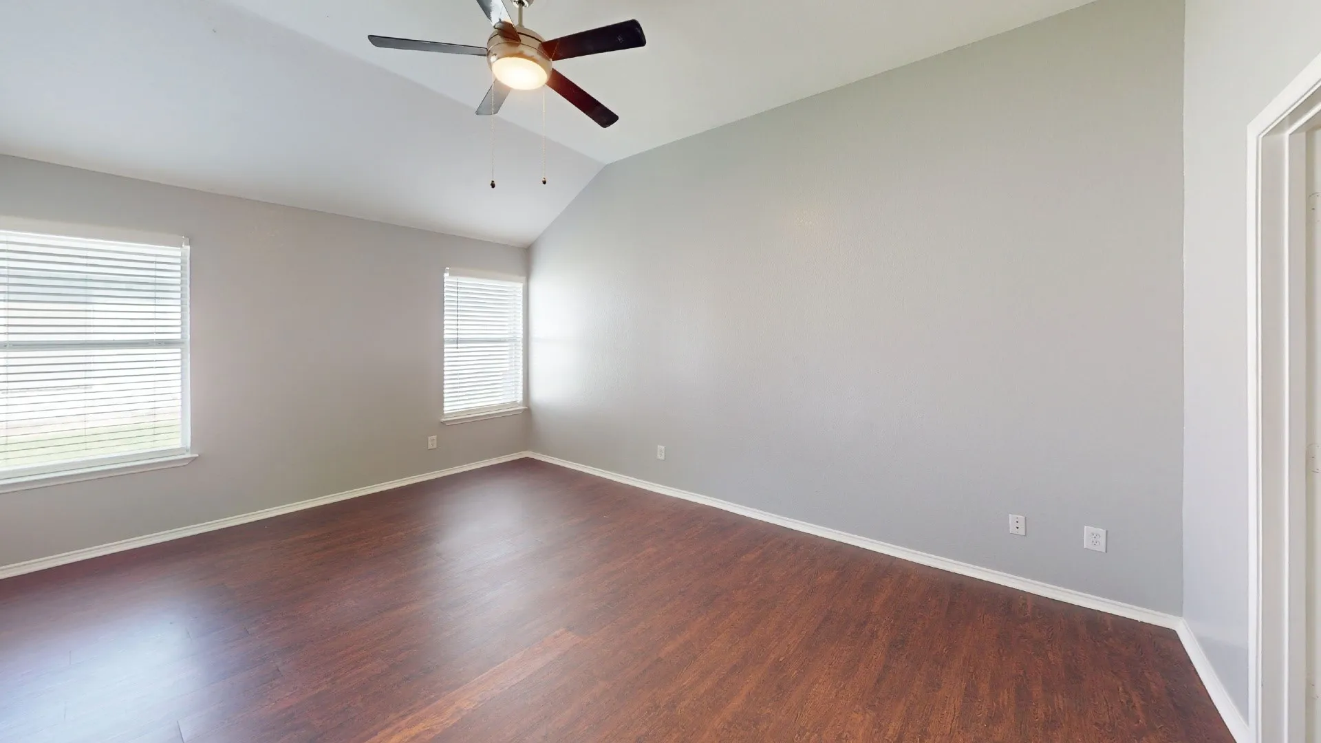 Vaulted ceiling with a ceiling fan, two windows with blinds, wood-finish flooring, and light gray walls