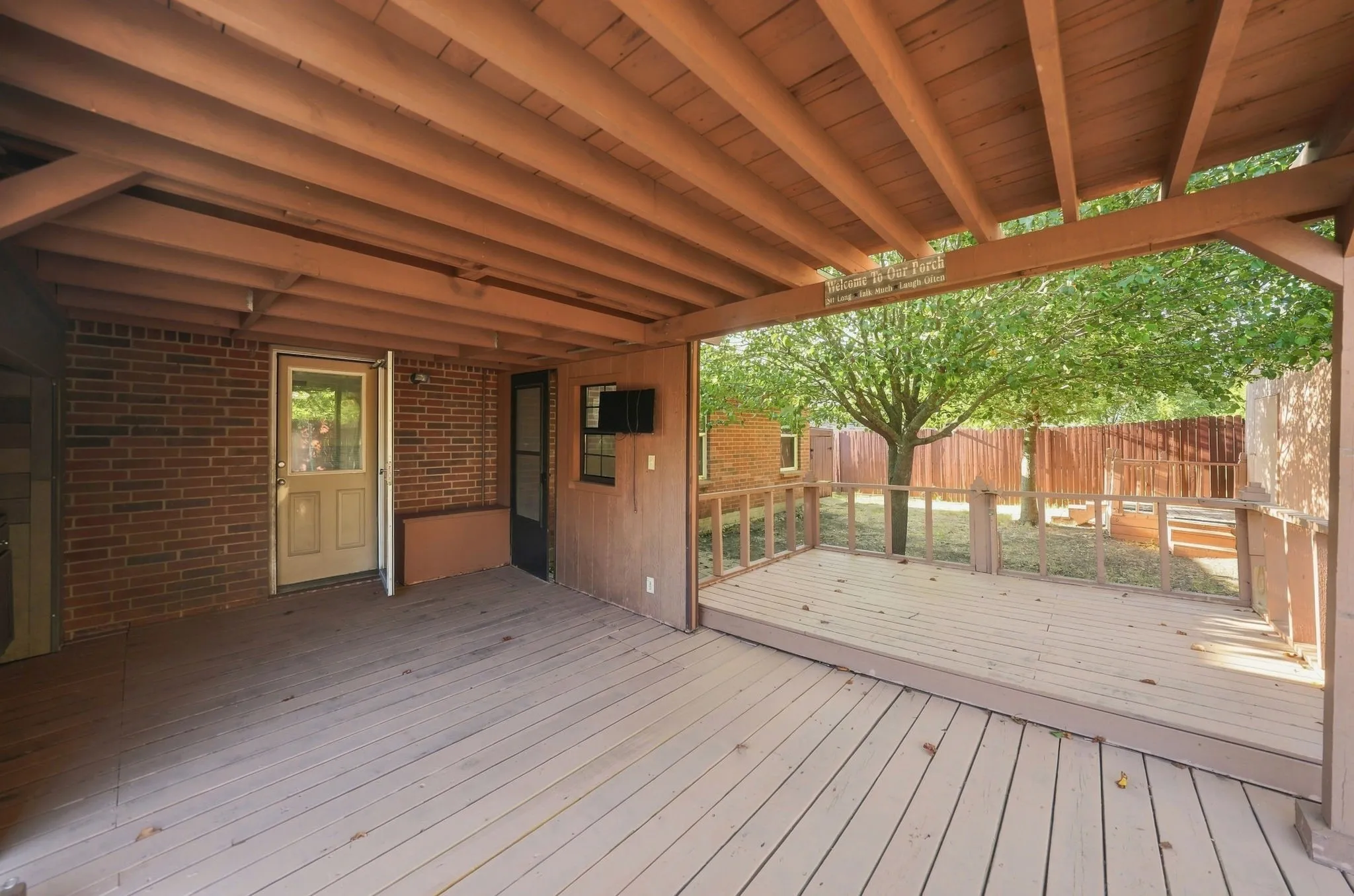Expansive covered deck with light-toned wood-finish flooring, exposed wood beam ceiling, brick exterior wall, and surrounding wooden privacy fence