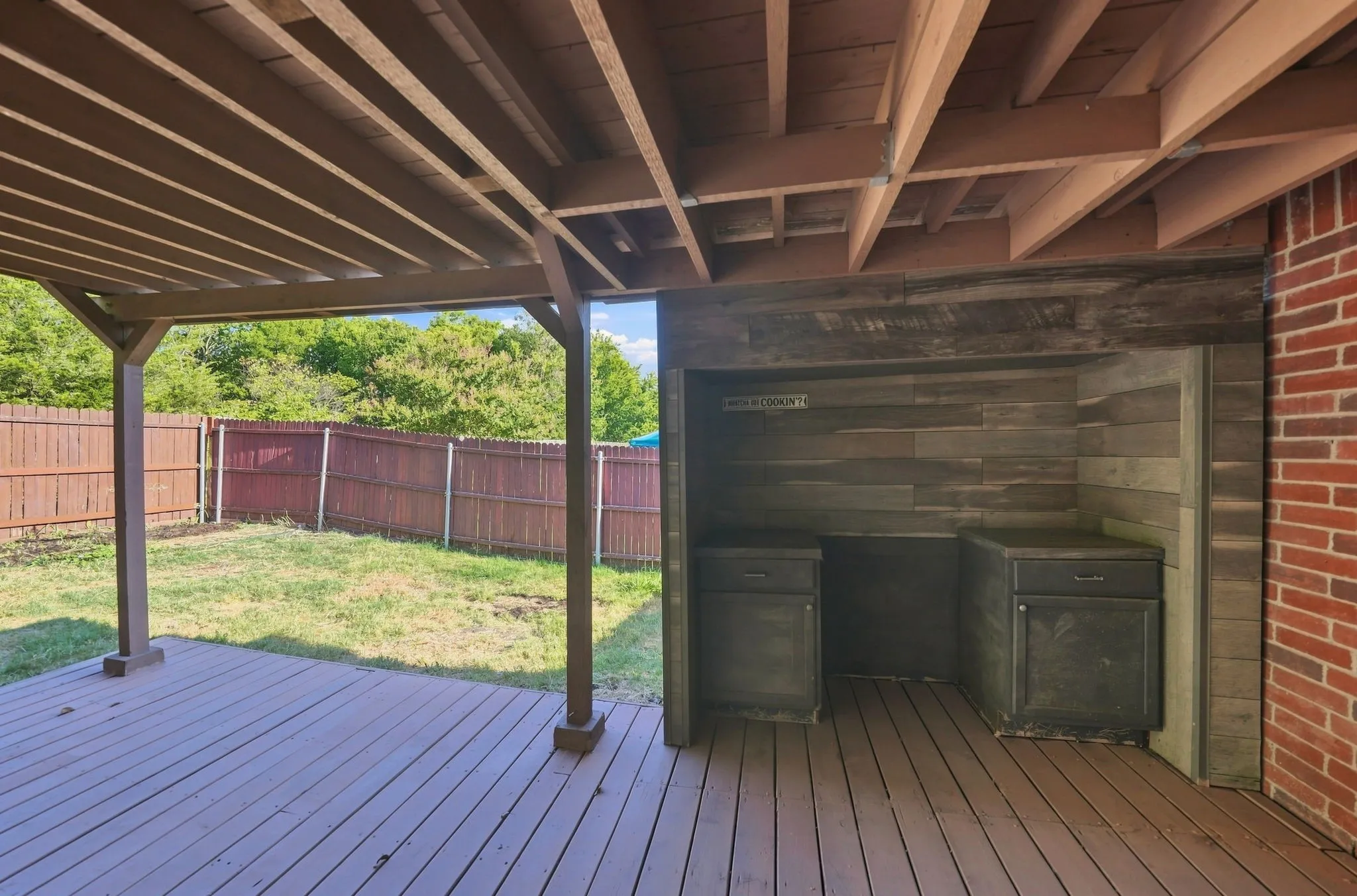Covered wood-finish deck with exposed structural beams, featuring a built-in outdoor kitchen area with cabinetry and a wood-finish accent wall