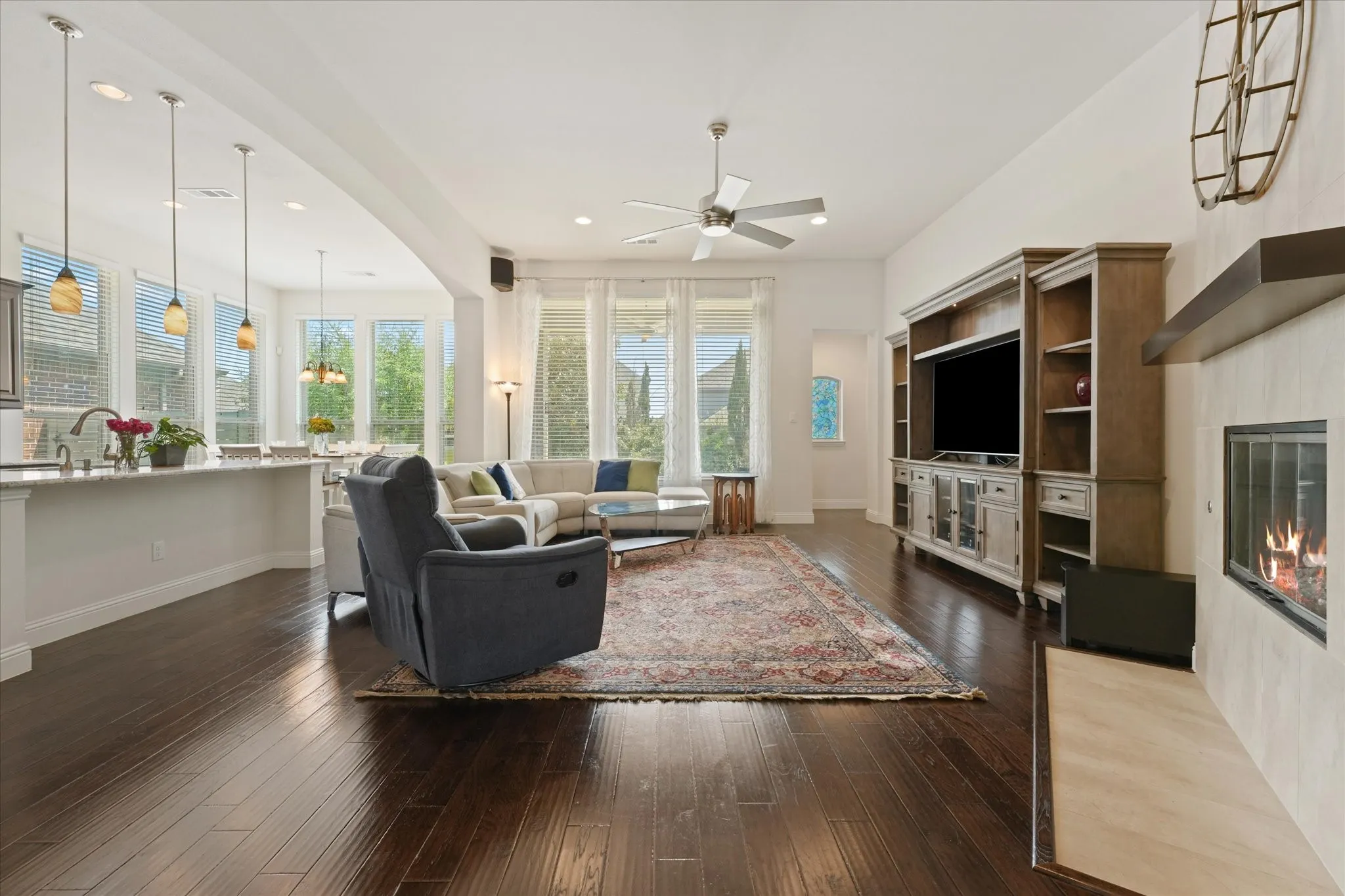 Open living room with a gas fireplace, engineered hardwood flooring, tall windows and direct views into the kitchen and breakfast area.