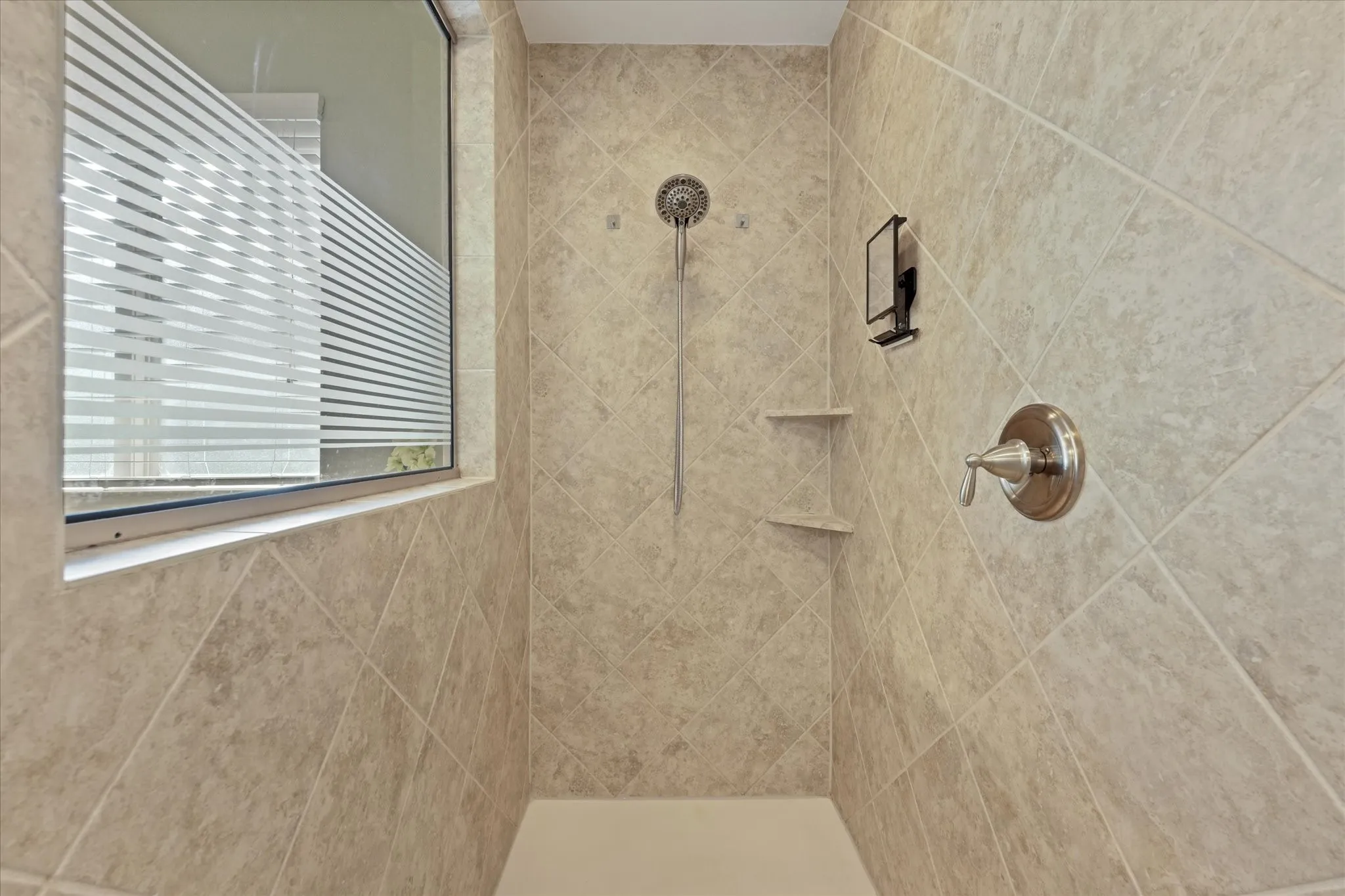 Oversized tiled walk-in shower with built-in shelving, handheld showerhead and private window for natural light.