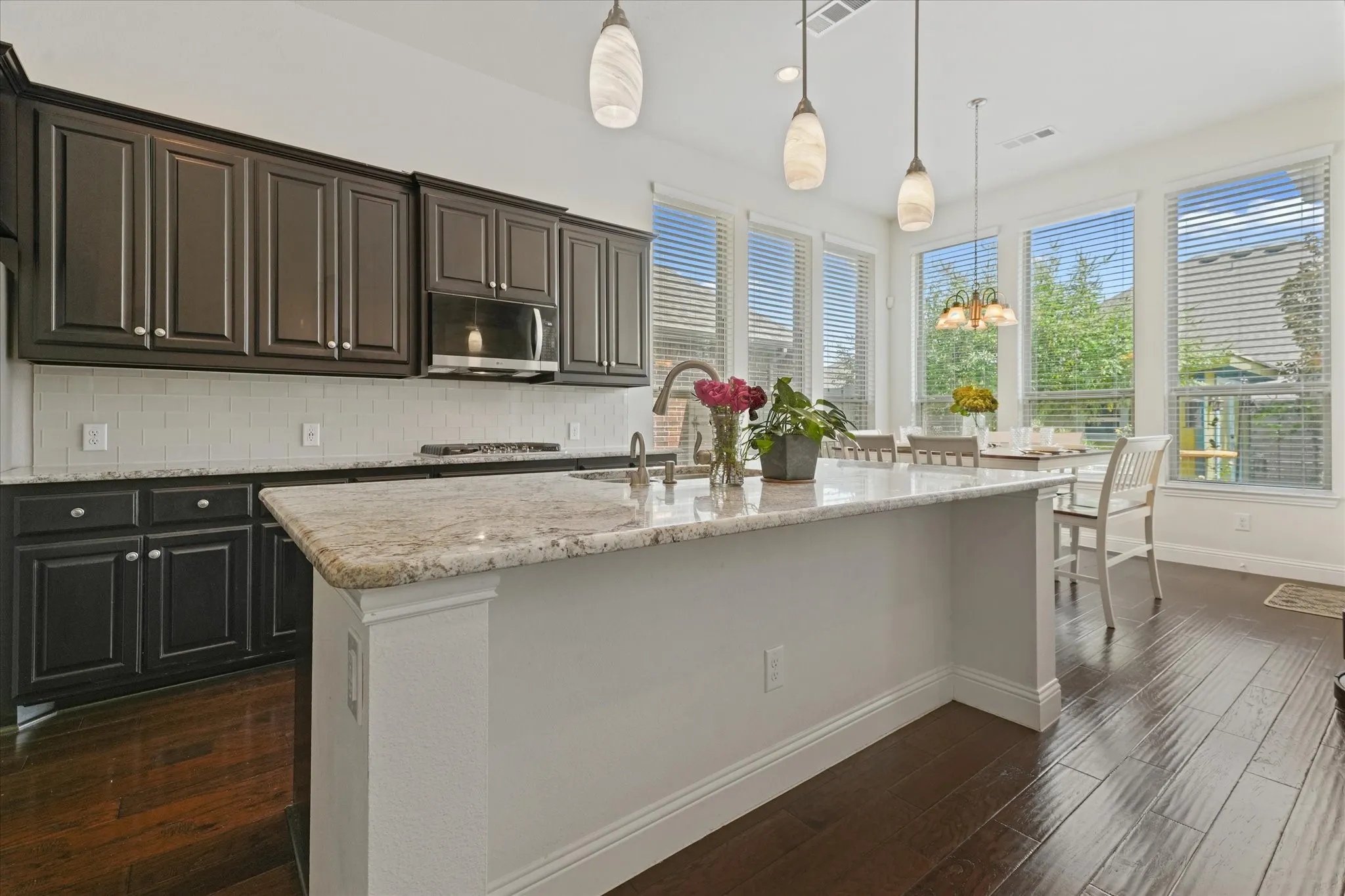 Open kitchen with a large granite island, dark cabinetry, pendant lighting and an adjoining breakfast area surrounded by windows.