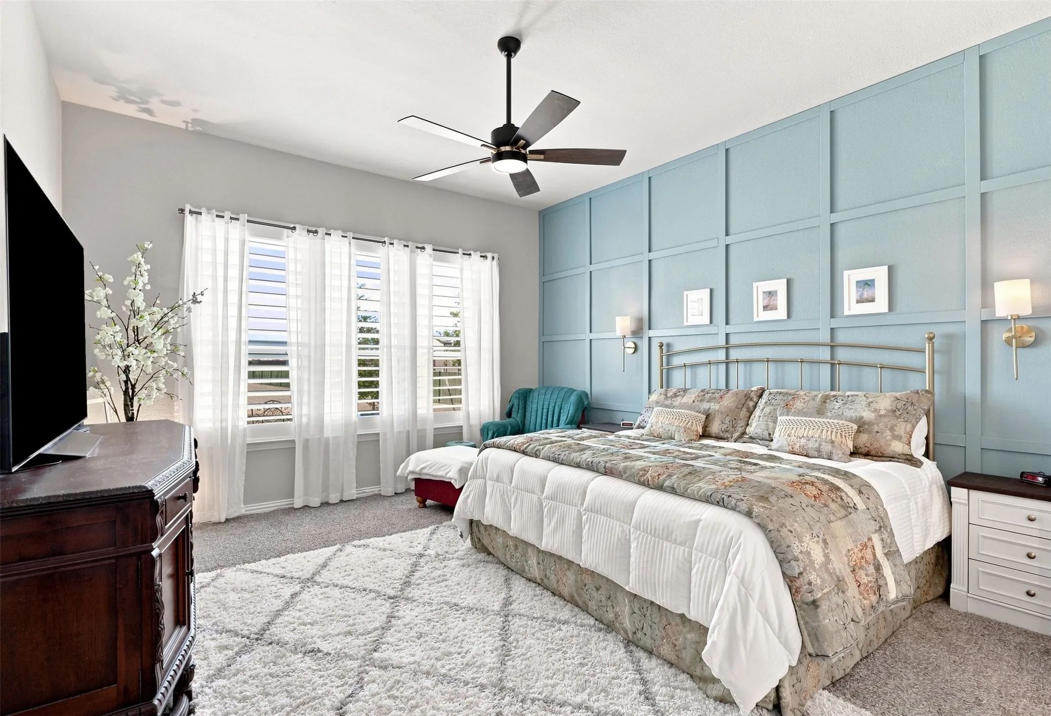 Primary Bedroom Suite featuring a decorative metal bed frame, dual nightstands with drawer storage, light-filtering shuttered doors opening to outdoor space, ceiling fan with dark finish, and a statement wall with blue-gray shiplap paneling and coordinating framed artwork