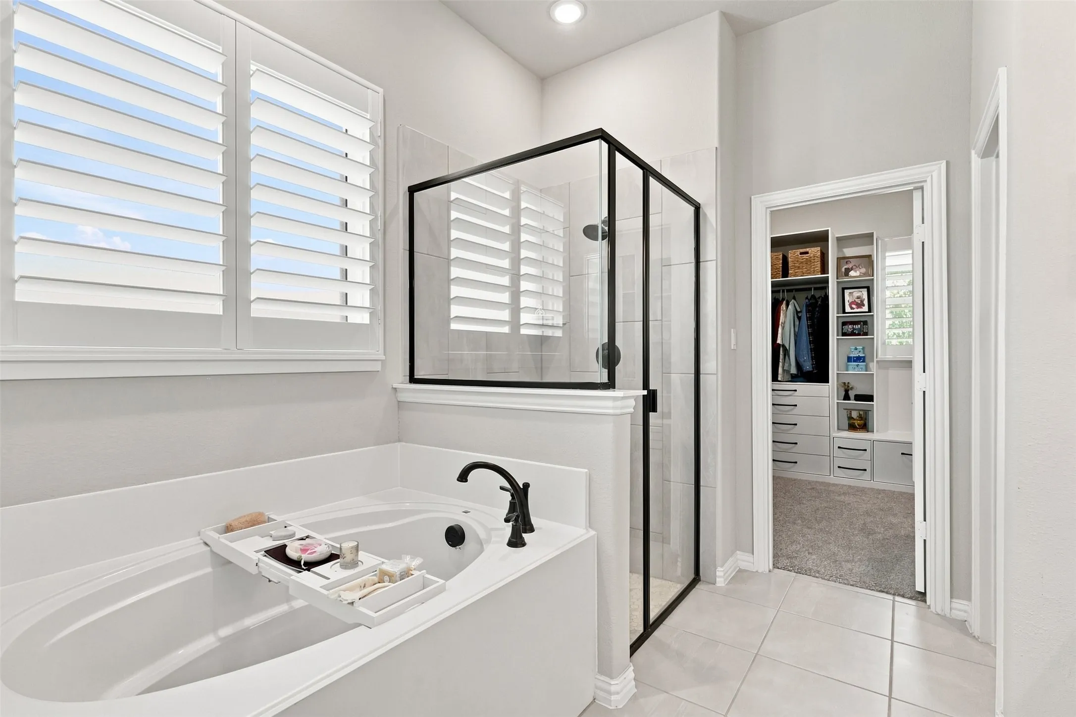 Spa-inspired primary bath featuring a freestanding soaking tub with matte black fixtures, a frameless glass-enclosed shower with black metal trim, porcelain tile flooring, plantation shutters, and abundant natural light from transom windows