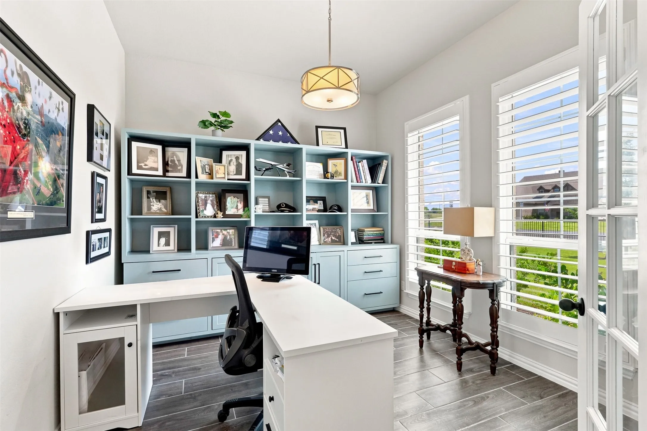 Contemporary home office featuring built-in cabinetry with open shelving and closed storage, a dual-desk workspace with white laminate countertops, polished concrete flooring, and abundant natural light from French doors and transom windows overlooking the outdoor landscape