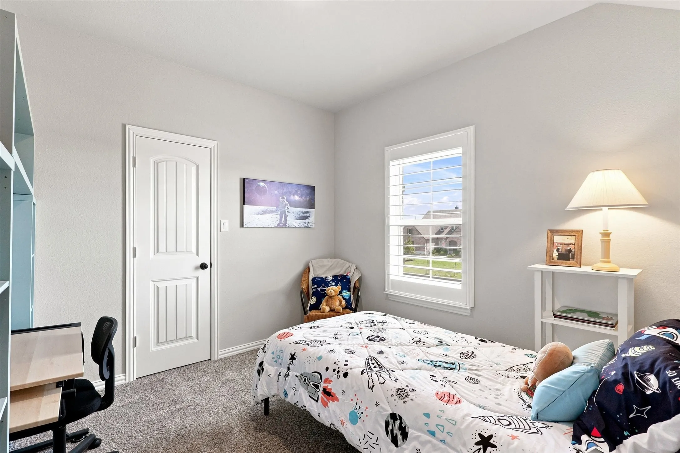 Bright secondary bedroom featuring carpet flooring, a large double-hung window with natural light, white six-panel closet door, and a bedside table with attached lighting fixture