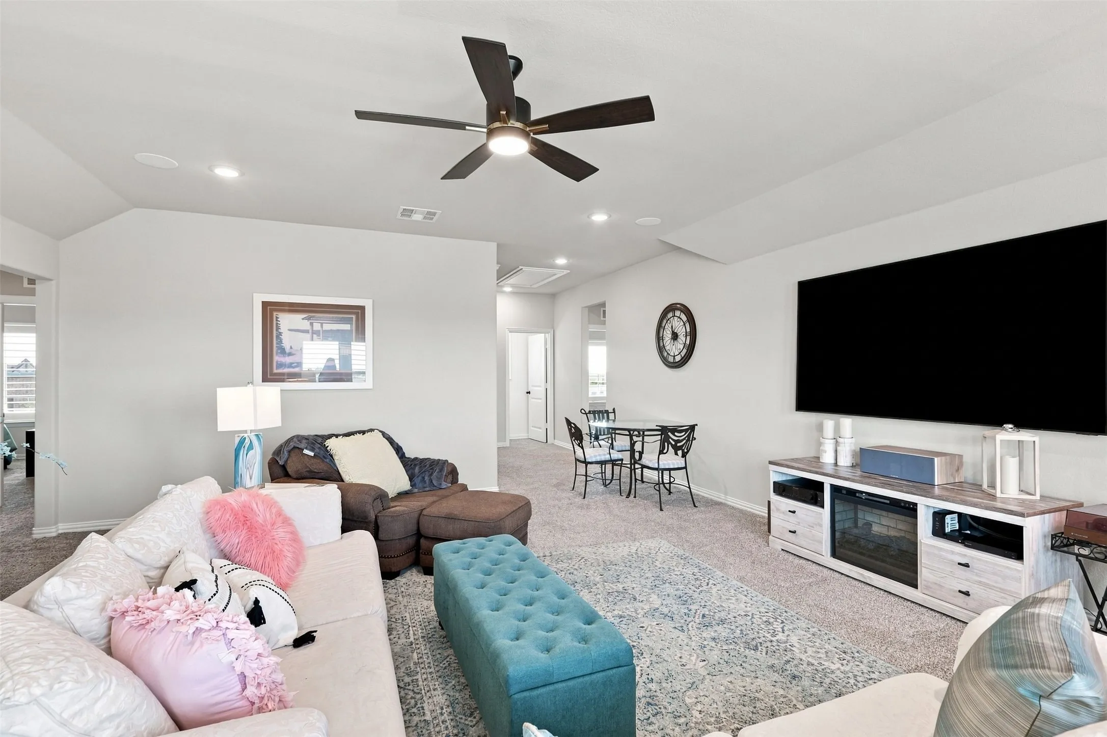 Spacious recreation room featuring recessed ceiling lighting, a ceiling fan with brushed metal finish, wall-mounted flat-screen television, built-in media console with shelving and storage, plush area rug, and direct access to adjacent spaces through multiple doorways