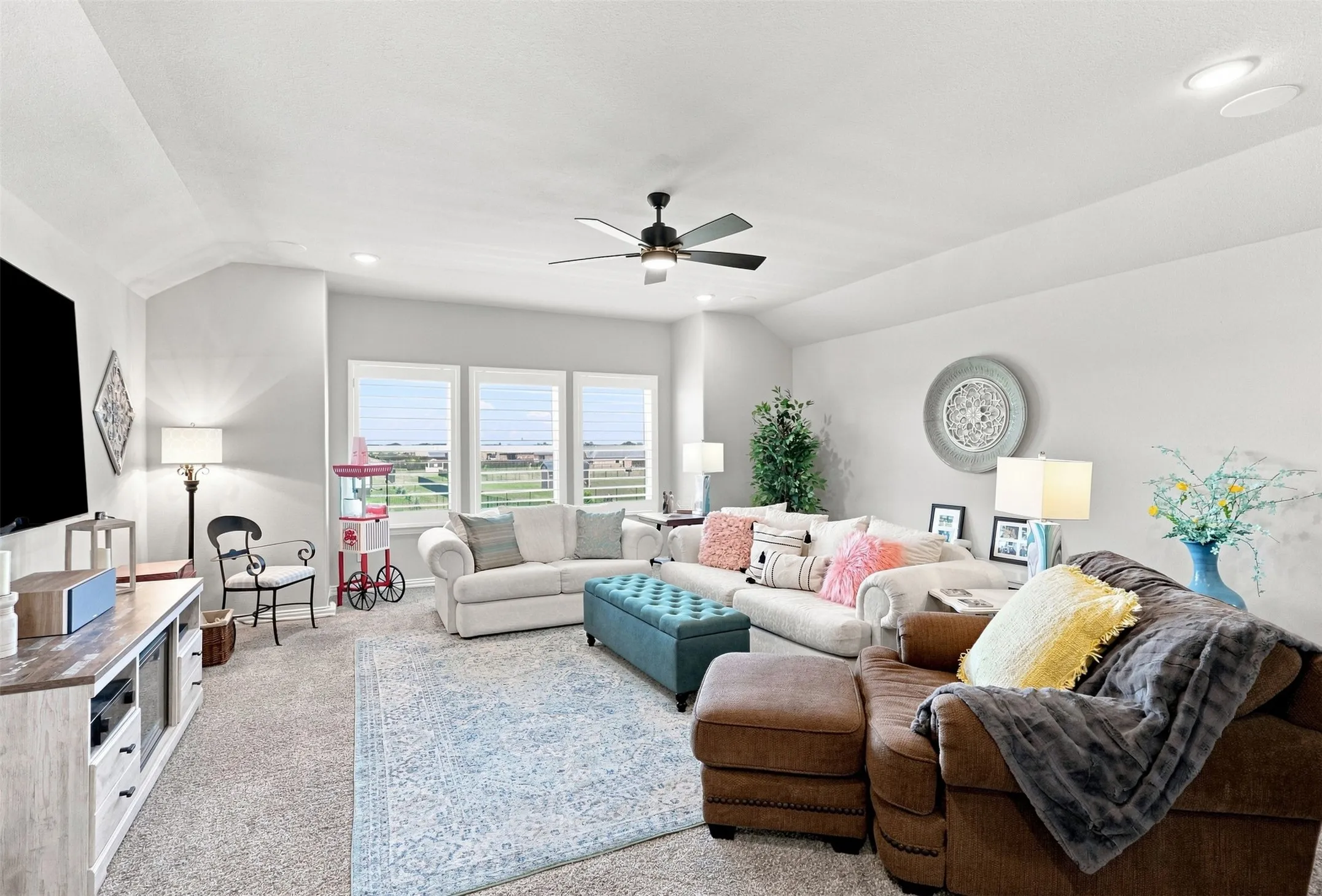 Bright and spacious living area featuring expansive windows with natural light, coffered ceiling detail, ceiling fan, and multiple seating zones anchored by a blue upholstered ottoman