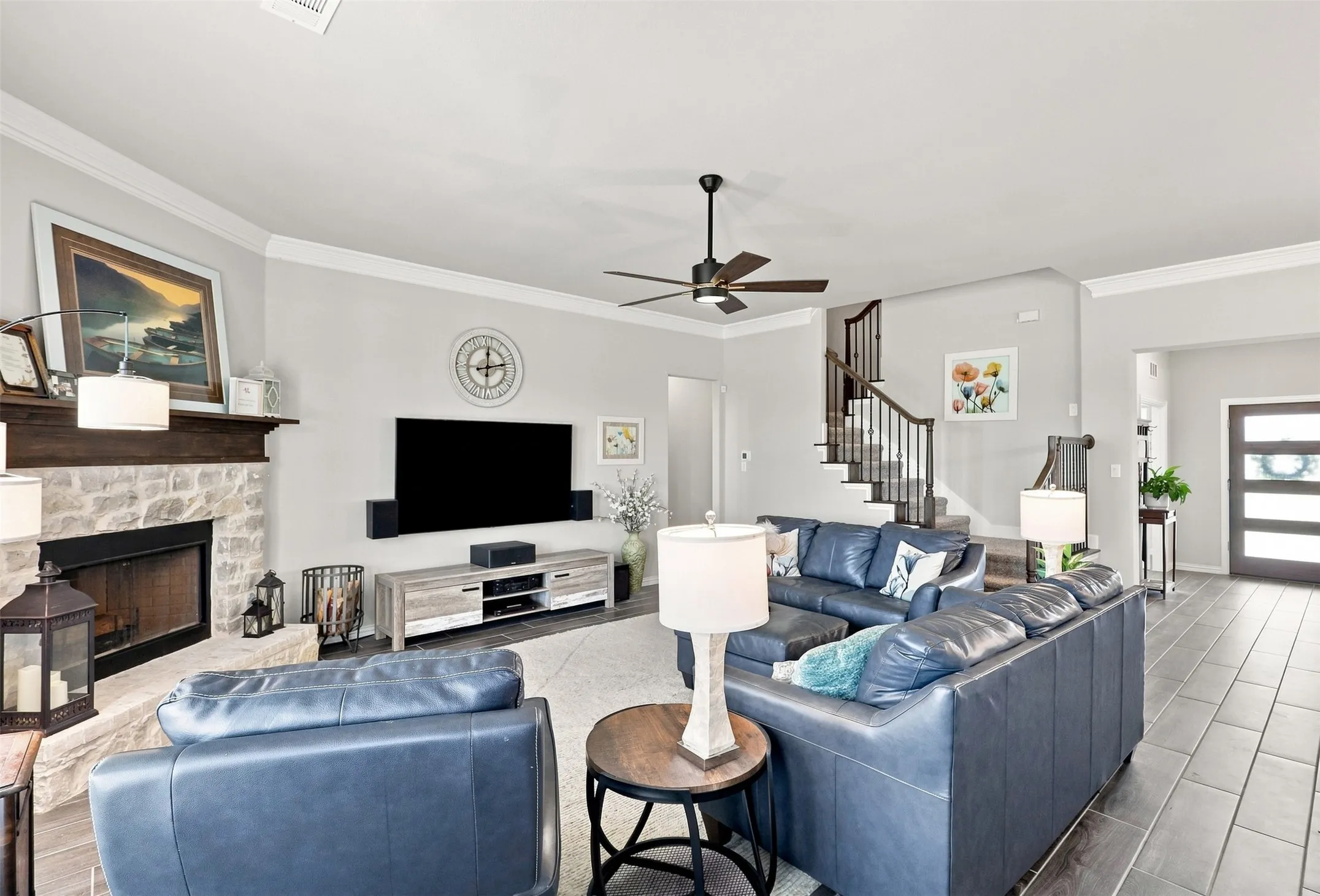Stone-faced fireplace with wood mantel, ceiling fan with integrated lighting, wood-finish flooring, staircase with dark wood handrail and balusters, and a modern entry door with frosted glass panels