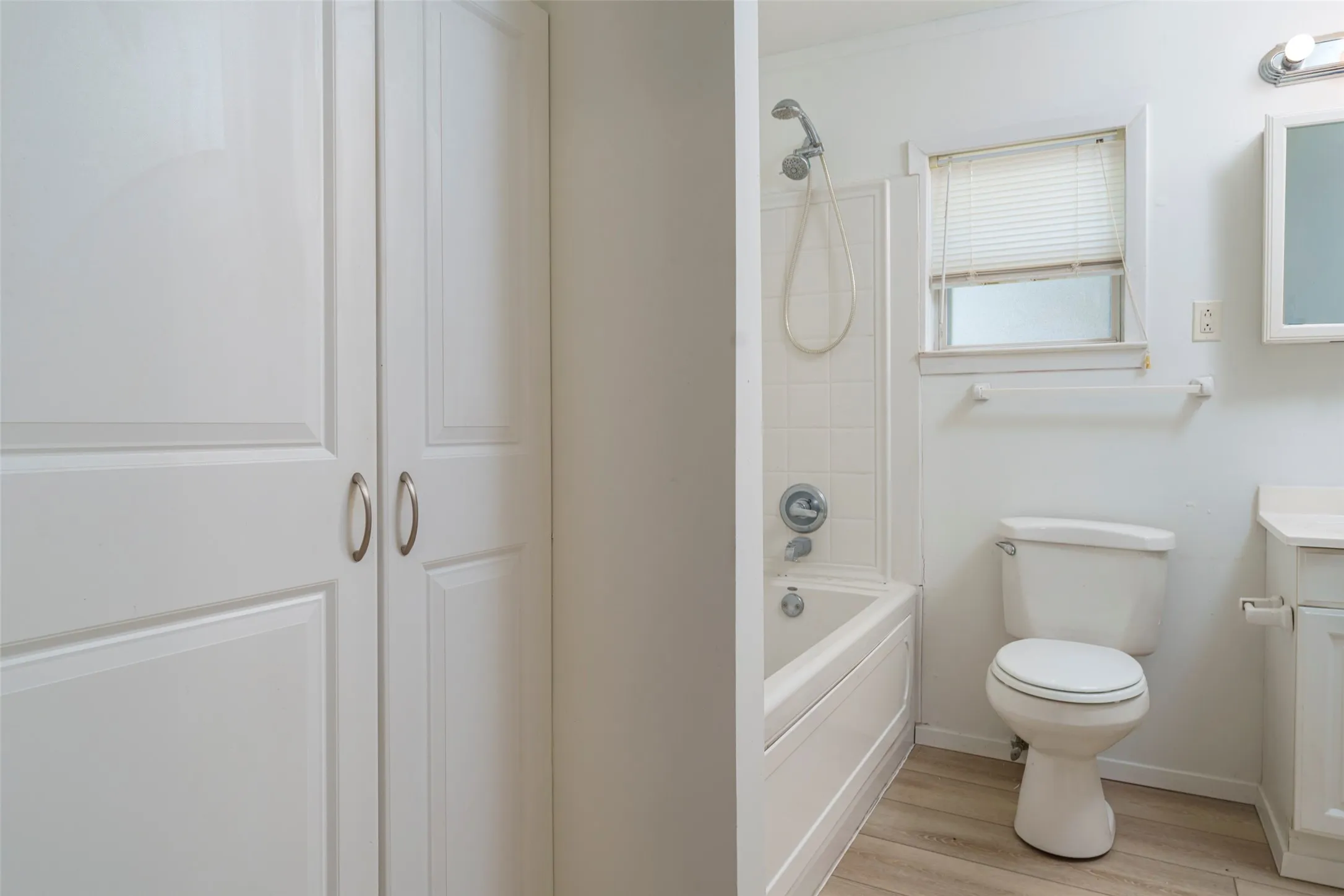 White paneled storage cabinetry, white bathtub with a shower, white toilet, single vanity with a white countertop, and wood-finish flooring