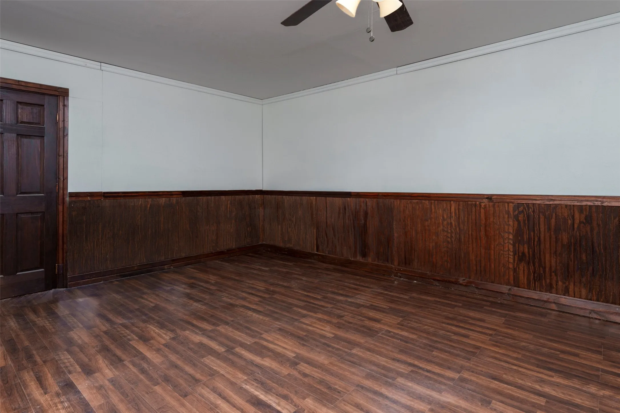 Wood-finish flooring, dark wood paneling, dark paneled door, ceiling fan with integrated lighting, and white crown molding