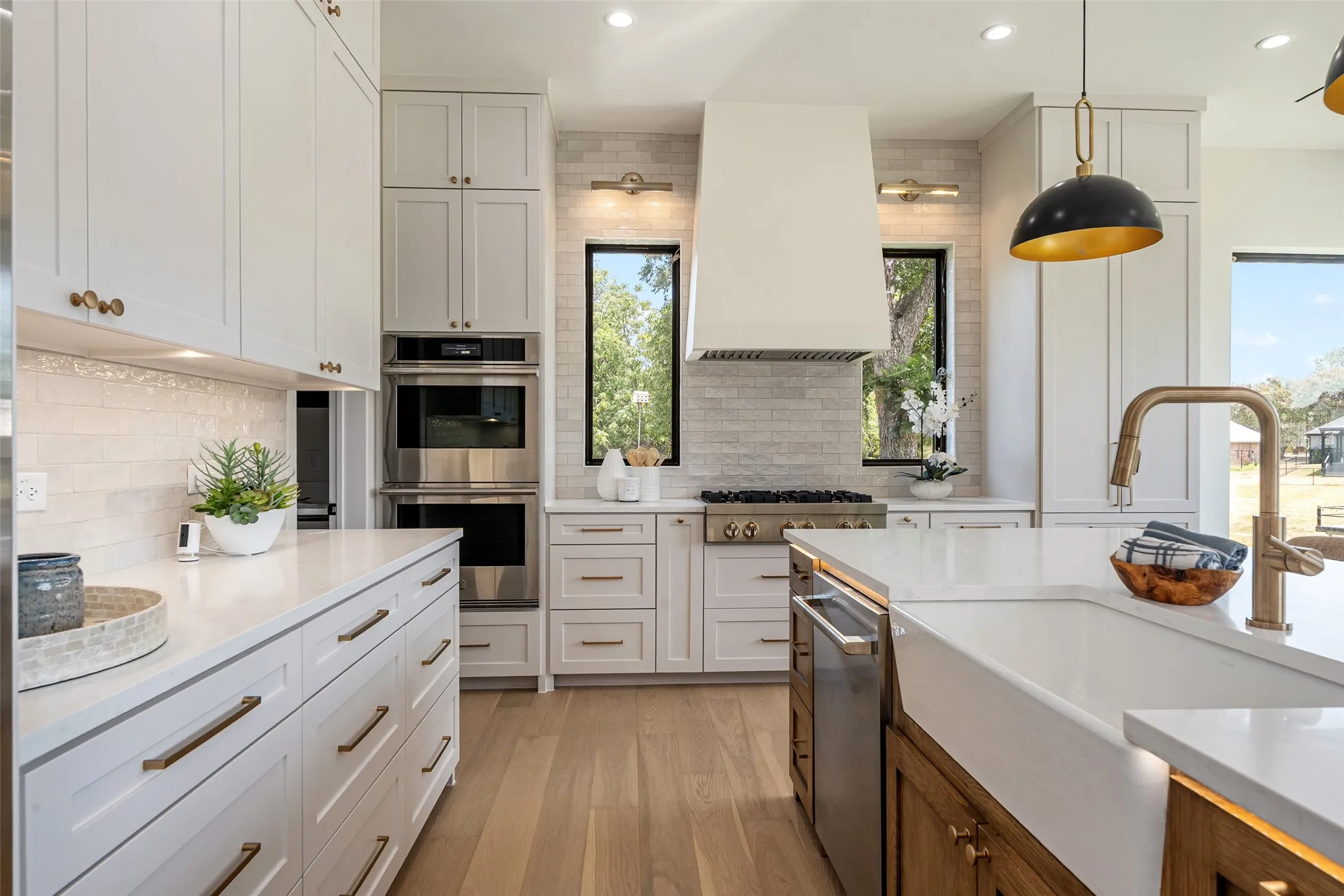 White shaker cabinetry with brass hardware, double wall ovens, wood-finish flooring, undermount farmhouse sink with gooseneck faucet, and an integrated dishwasher