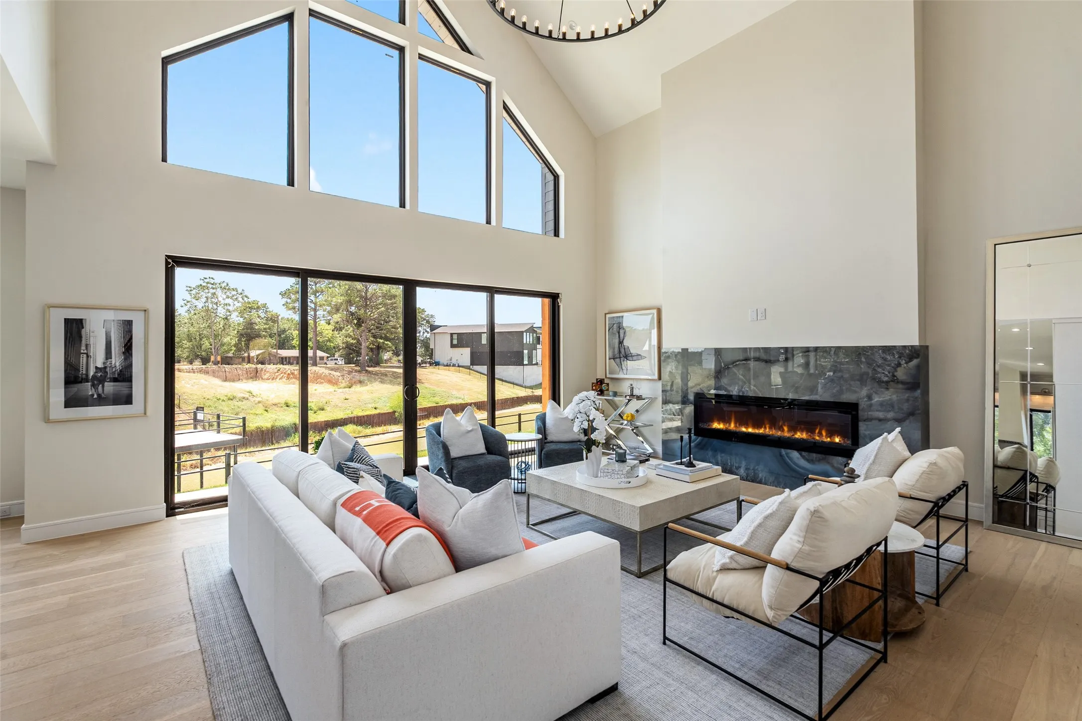 High vaulted ceiling with upper-level transom windows, black-framed sliding glass doors, a modern electric fireplace with a stone surround, and light wood-finish flooring