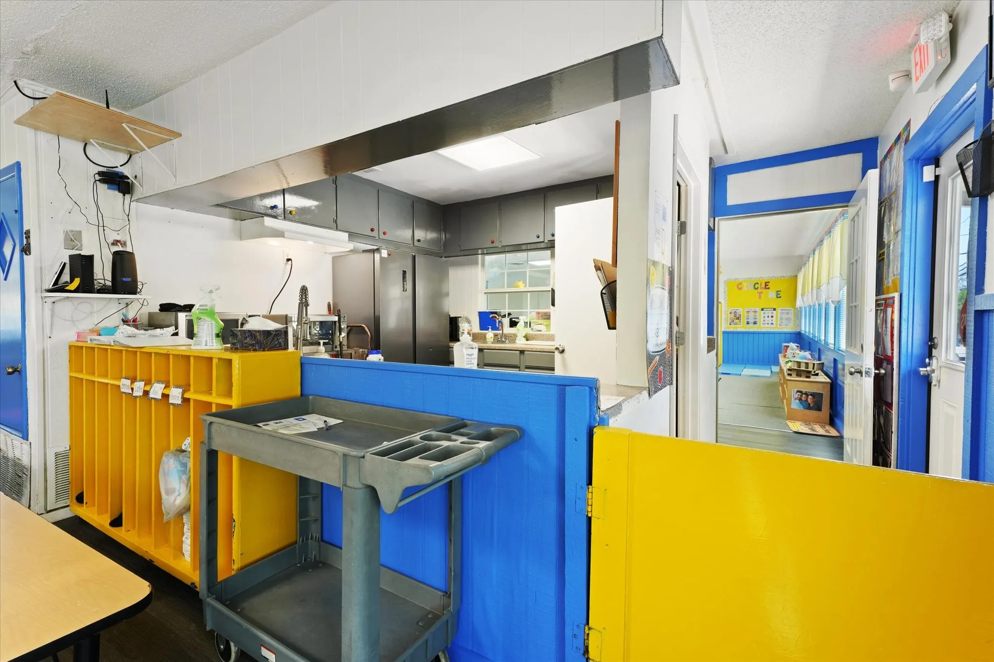 Modern commercial kitchen featuring bold blue and yellow cabinetry, stainless steel countertops and prep surfaces, professional-grade cooking equipment, ample storage solutions, and bright overhead lighting with contemporary design elements throughout