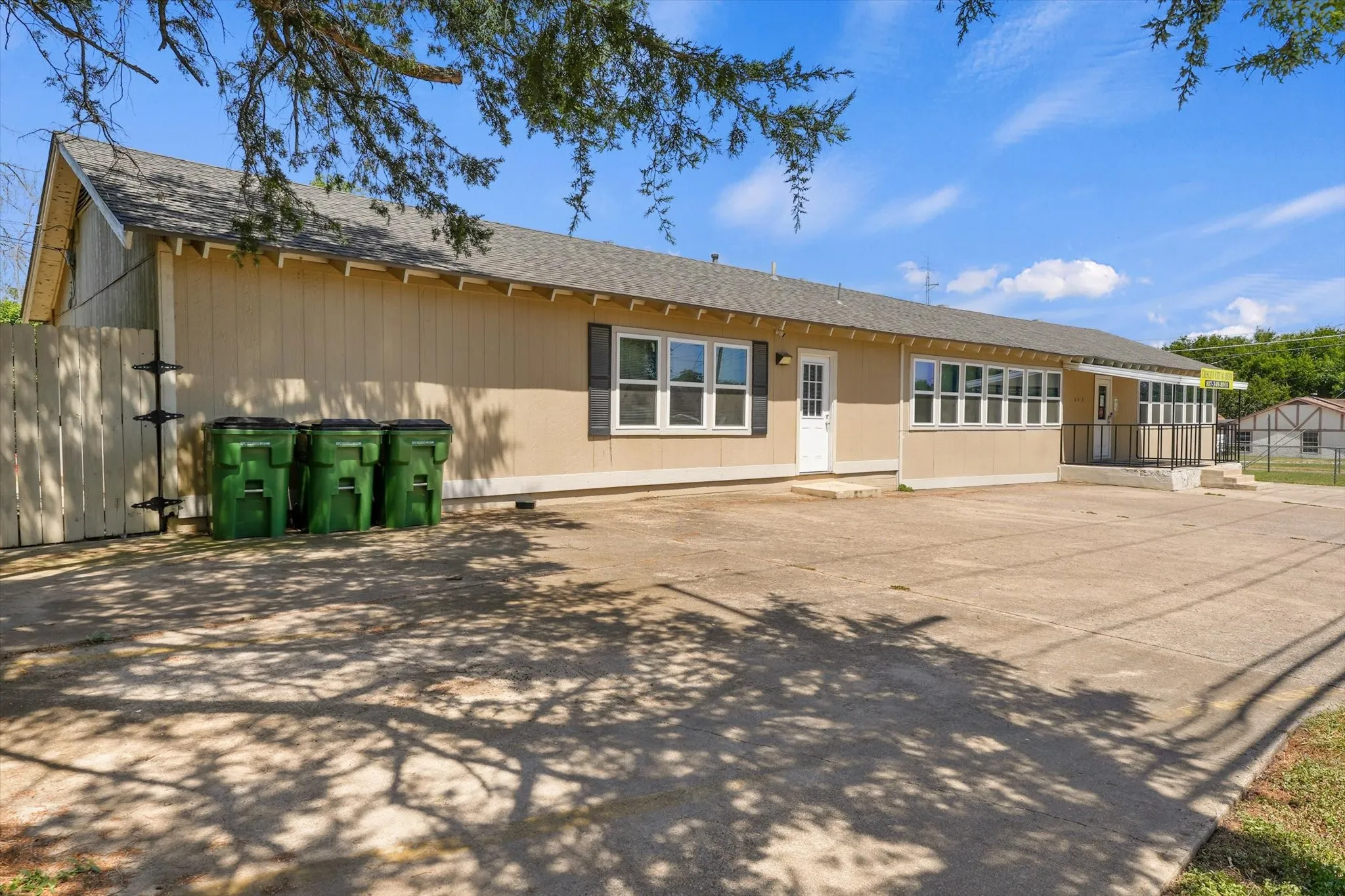 Spacious single-story commercial or institutional building featuring a tan exterior with multiple window banks, concrete patio area, and mature tree coverage