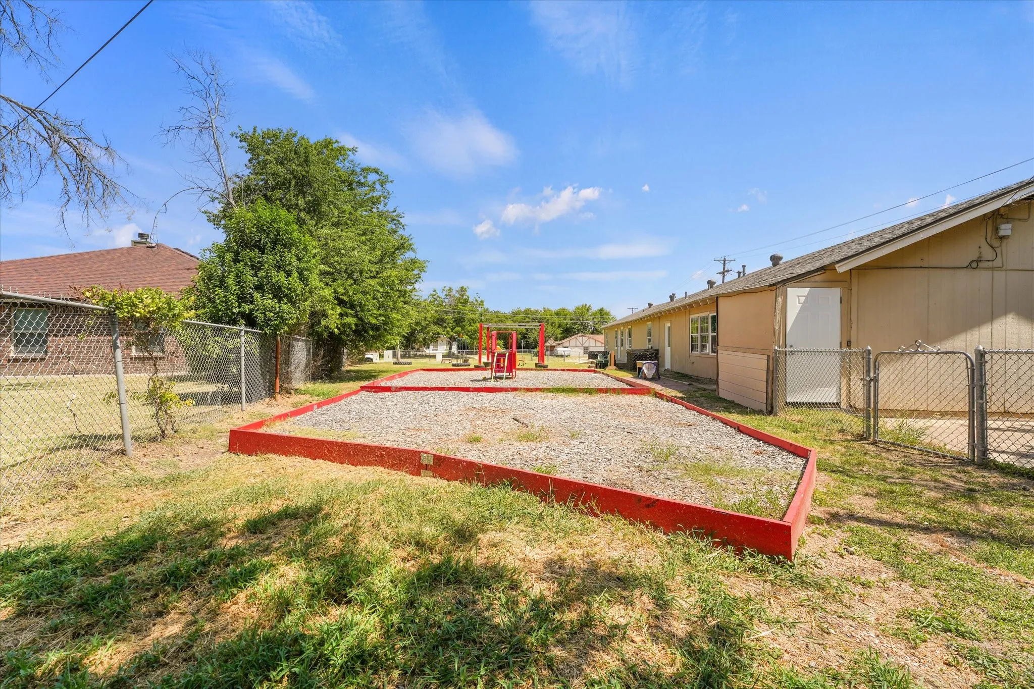 Outdoor recreational area featuring a red-bordered, gravel-filled play space, a chain-link fence perimeter, and mature trees in the background