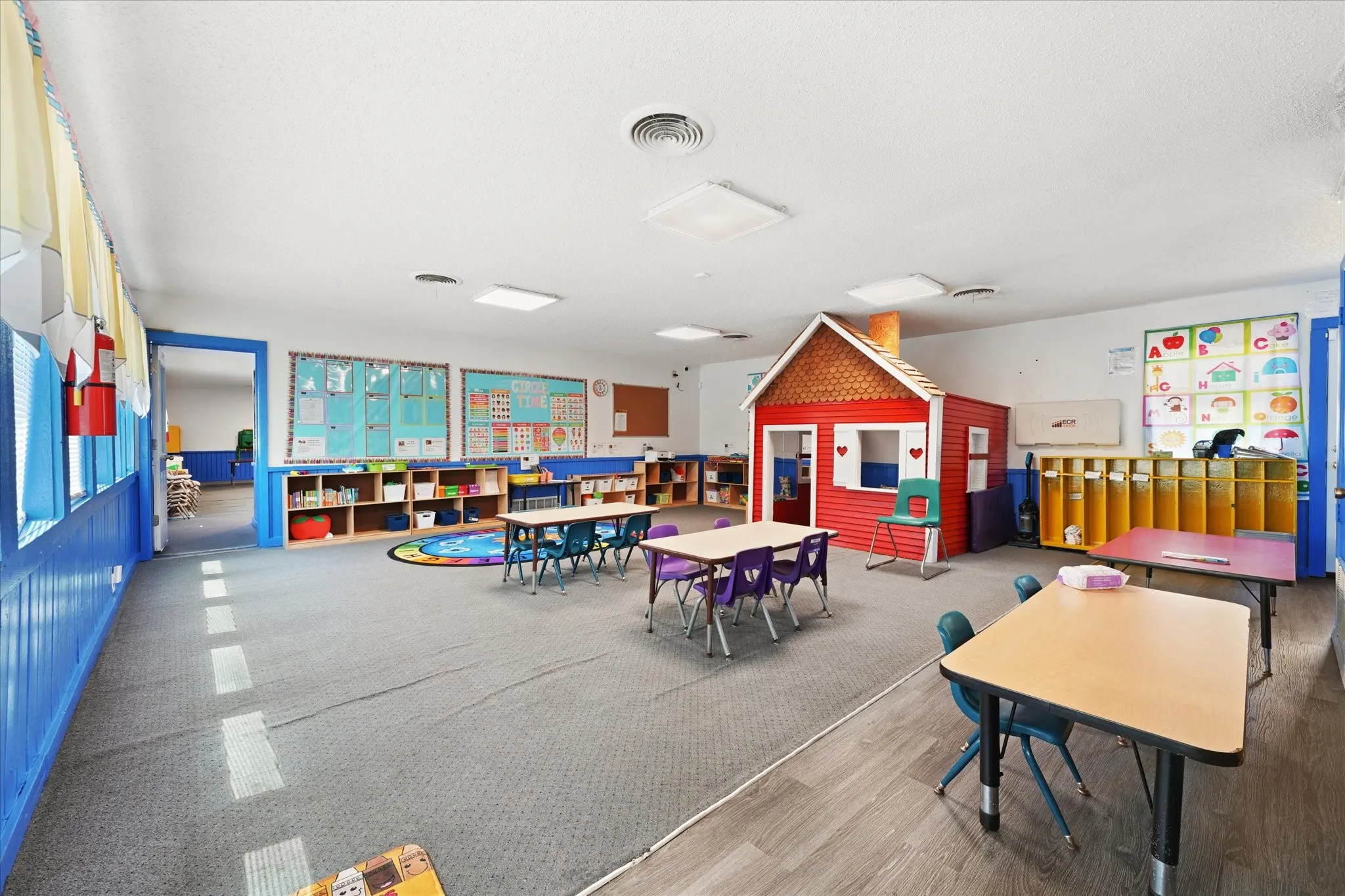 Bright, open learning environment featuring flexible furniture arrangements, built-in storage cabinetry along perimeter walls, recessed ceiling lighting, wood-finish flooring, and a custom play structure with red and blue accents