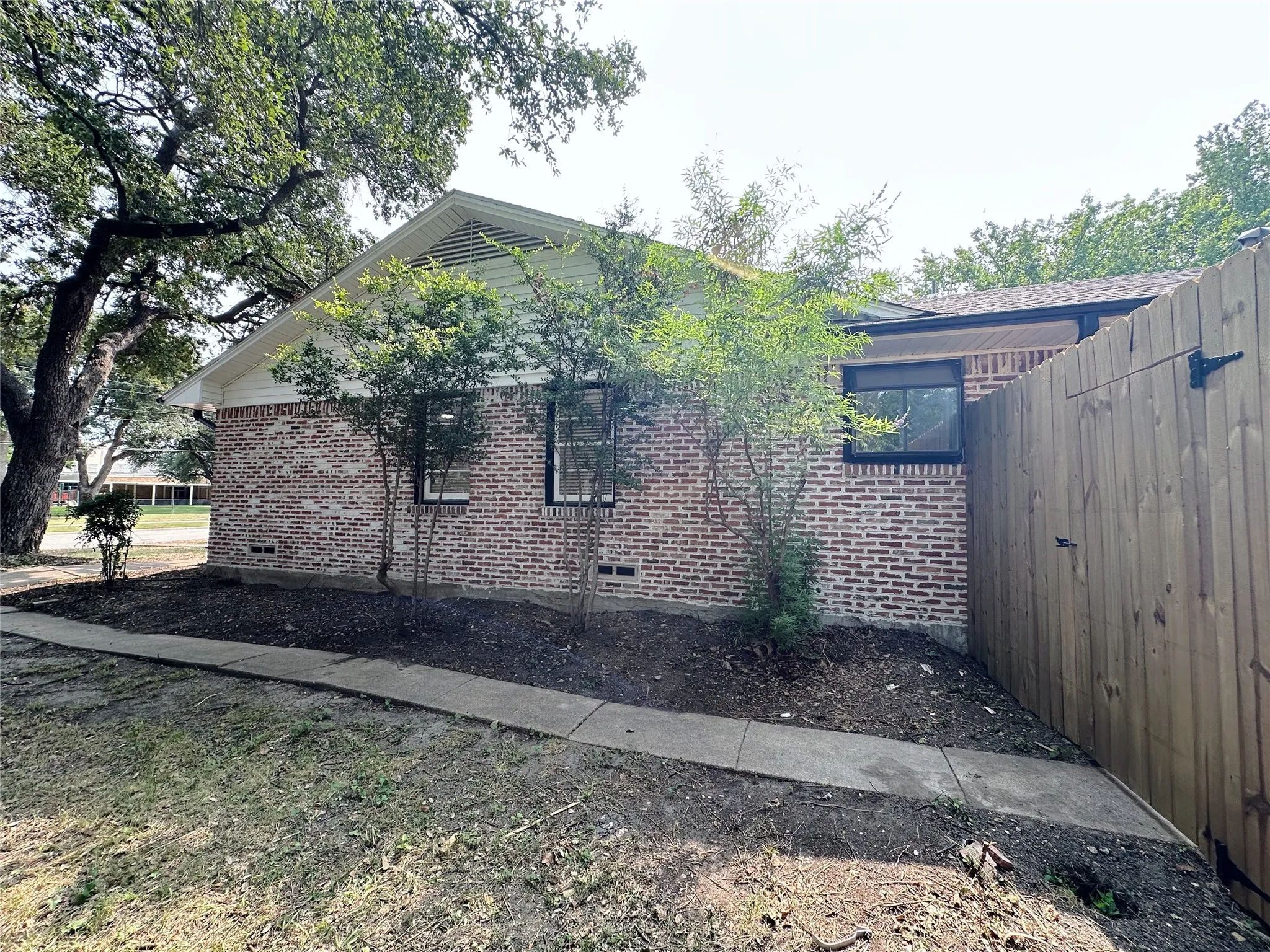 Brick exterior with a gabled roofline, mature trees, and a privacy fence