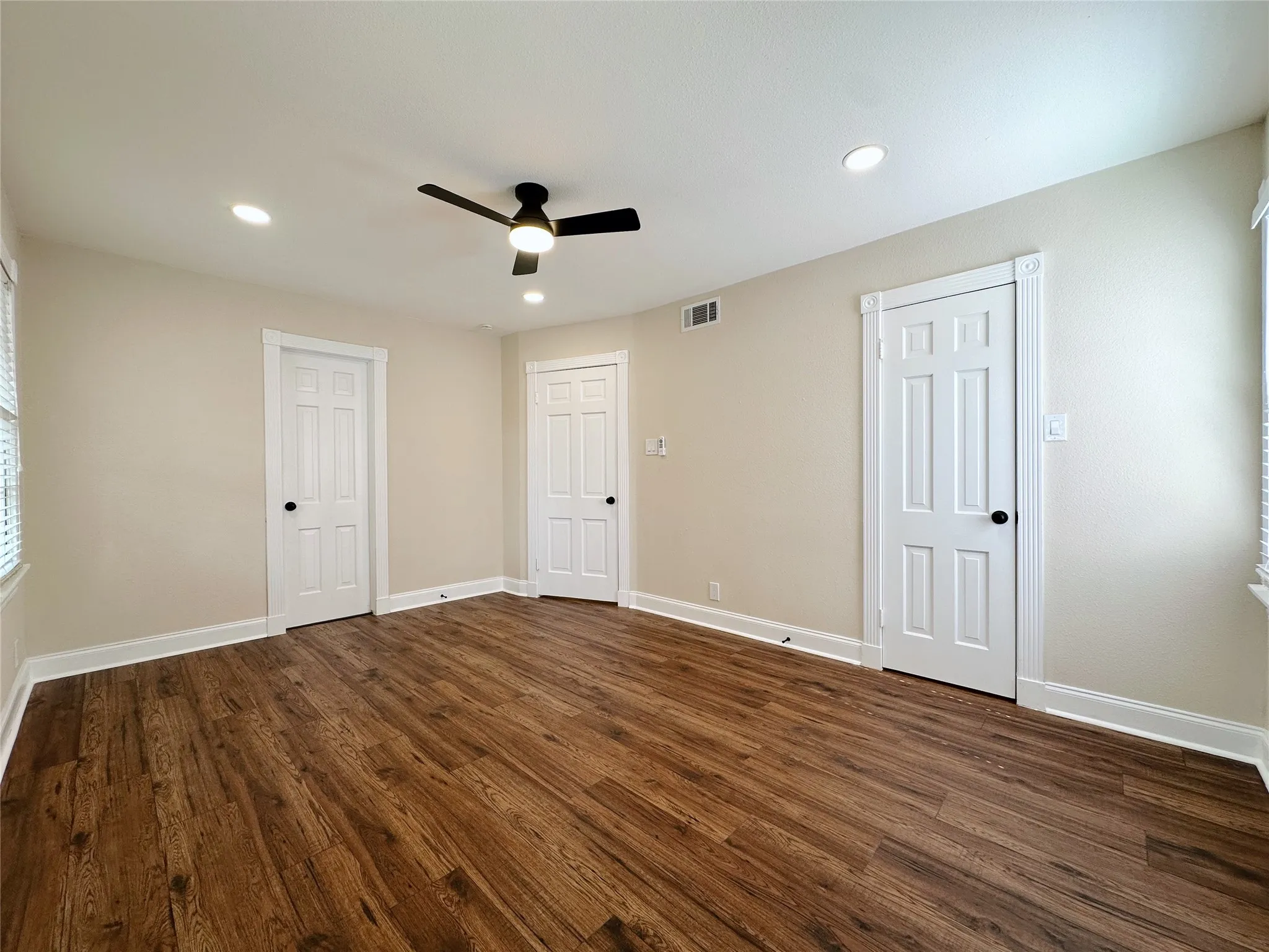 Spacious room featuring three white six-panel doors, warm neutral walls, ceiling fan with integrated lighting, and rich wood-finish flooring throughout