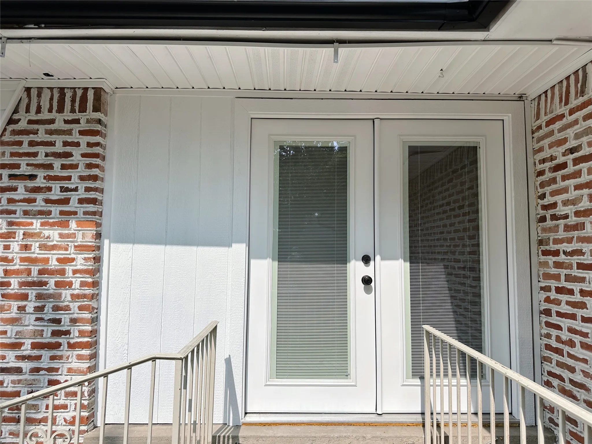Red brick exterior, white vertical panel siding, white double French doors with integrated blinds, and a white metal handrail