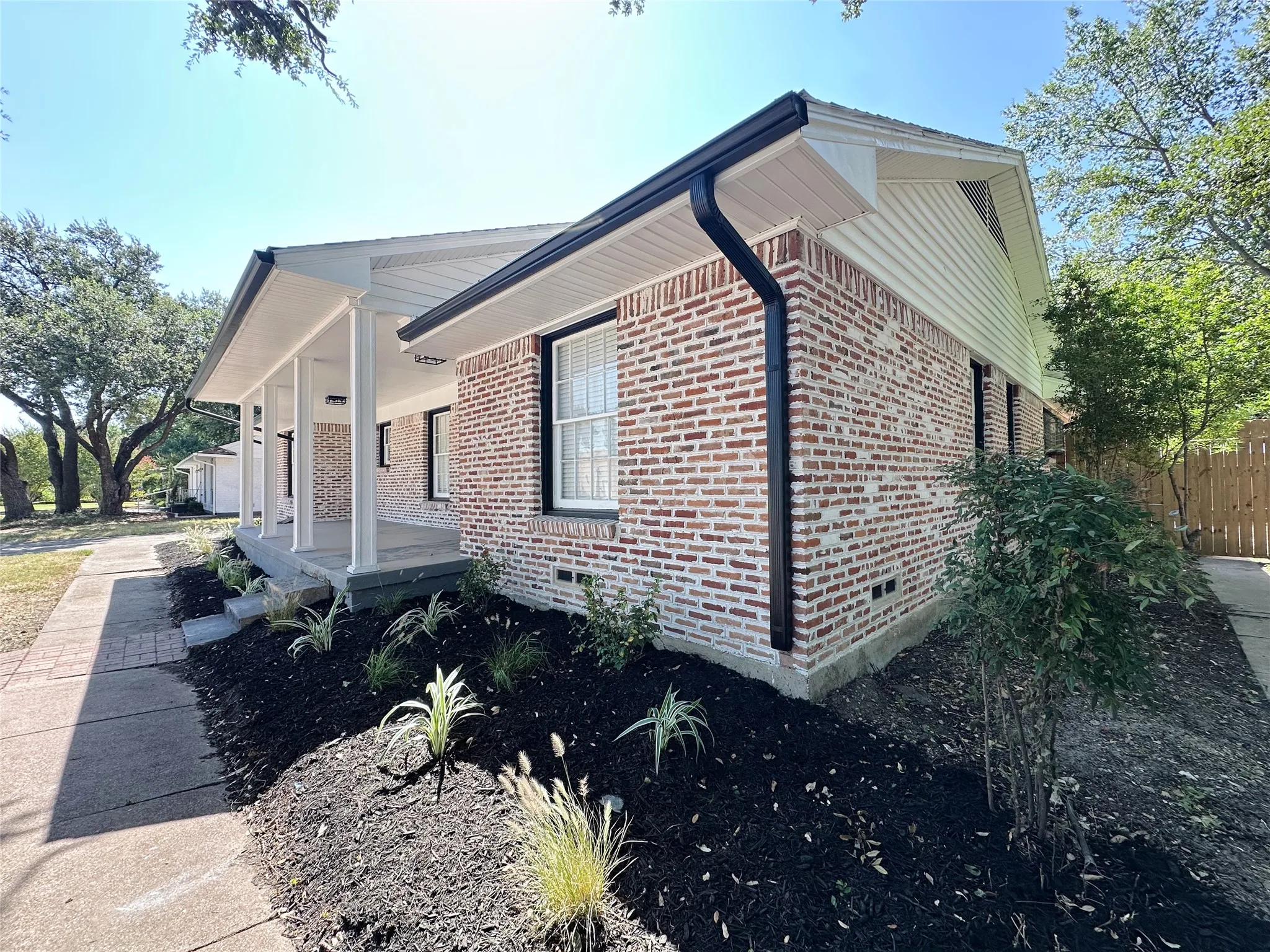 Brick exterior with white siding accents, covered front porch with multiple columns, dark gutters, and landscaped front yard with planting beds