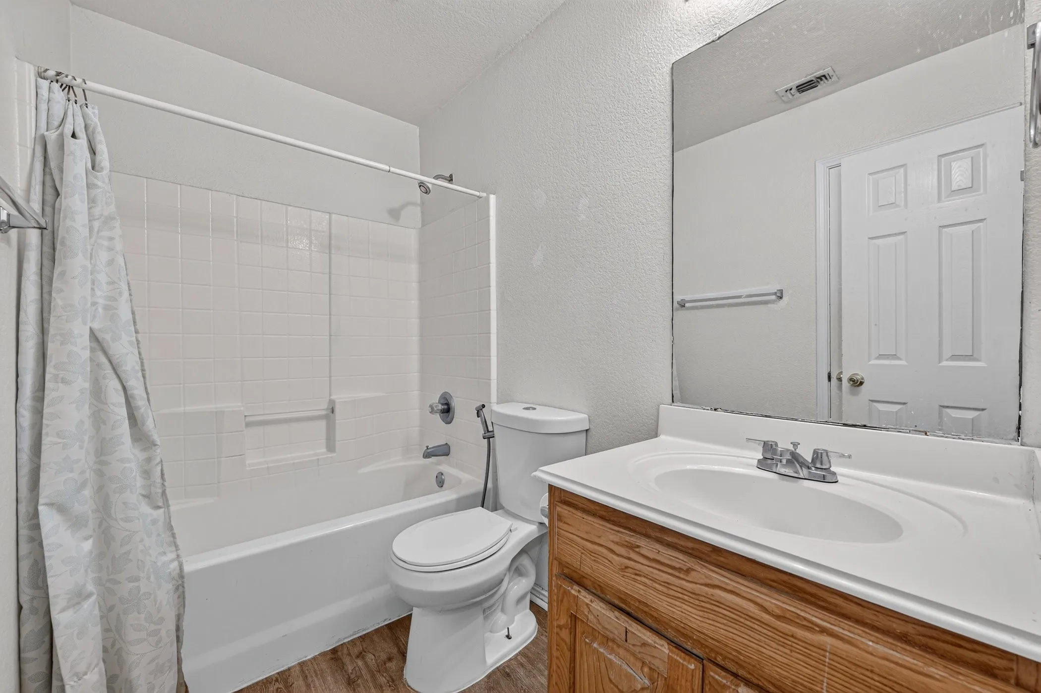 Bathroom featuring a wood-finish vanity with an integrated sink, a large wall-mounted mirror, white subway tile shower surround, and wood-finish flooring