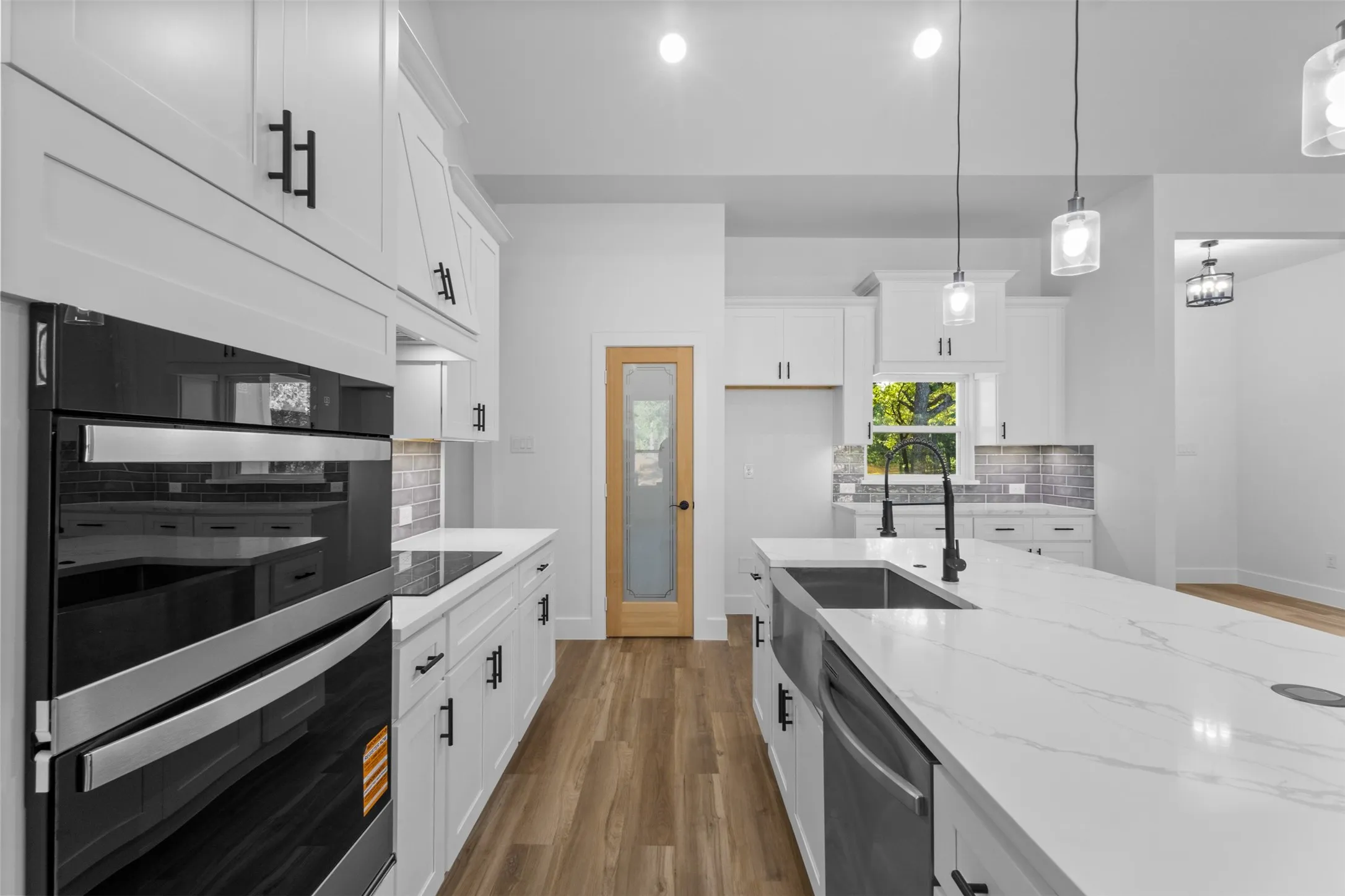 Modern kitchen featuring white cabinetry with integrated hardware, quartz countertops, a waterfall-edge island with undermount sink, stainless steel appliances including dual wall ovens, pendant lighting, wood-finish flooring, and abundant natural light from multiple windows