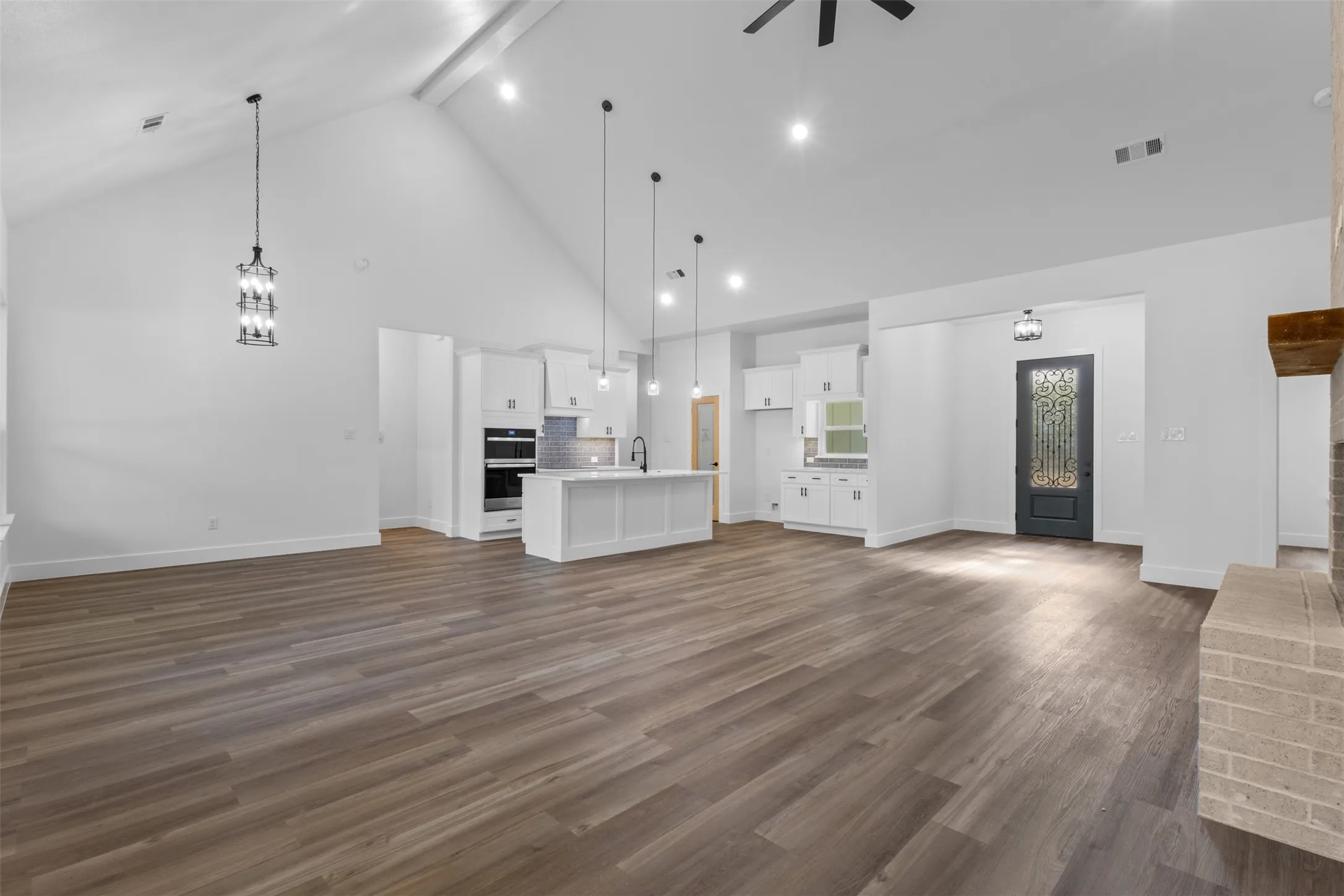 Open-concept living space featuring soaring ceilings with pendant lighting, wide-plank wood-finish flooring throughout, and a central kitchen island with white cabinetry