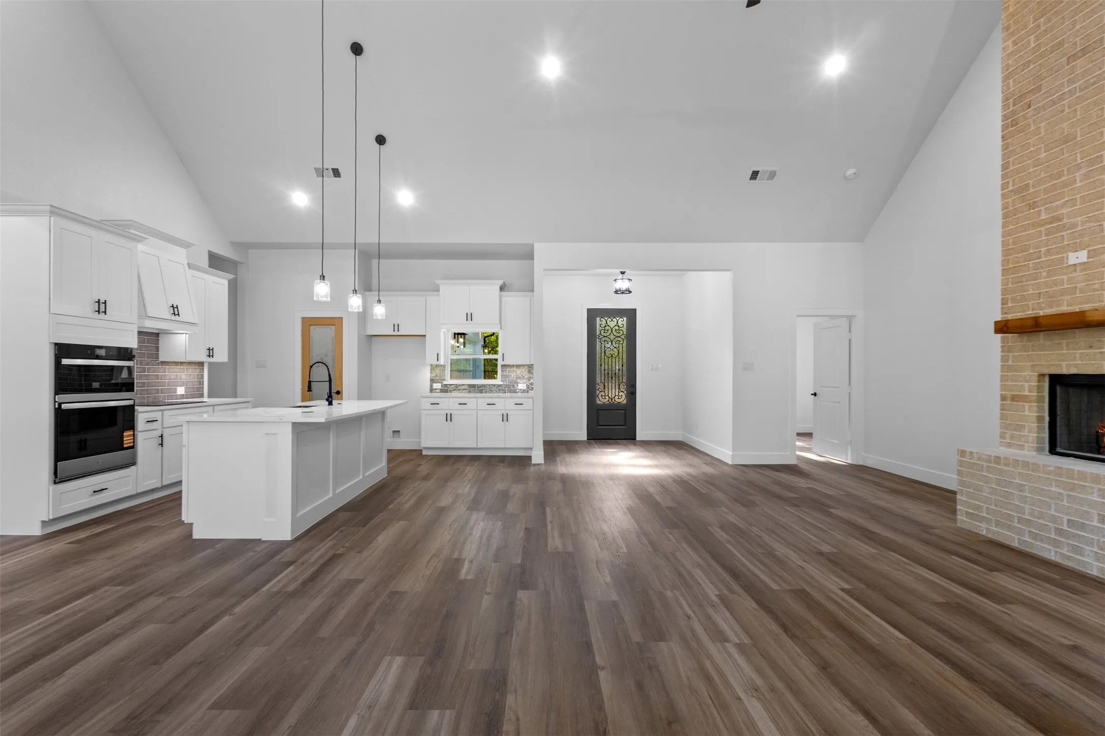 Open-concept kitchen and living area featuring white cabinetry with hardware accents, stainless steel built-in appliances, a large island with pendant lighting, wood-finish flooring throughout, and recessed ceiling lighting