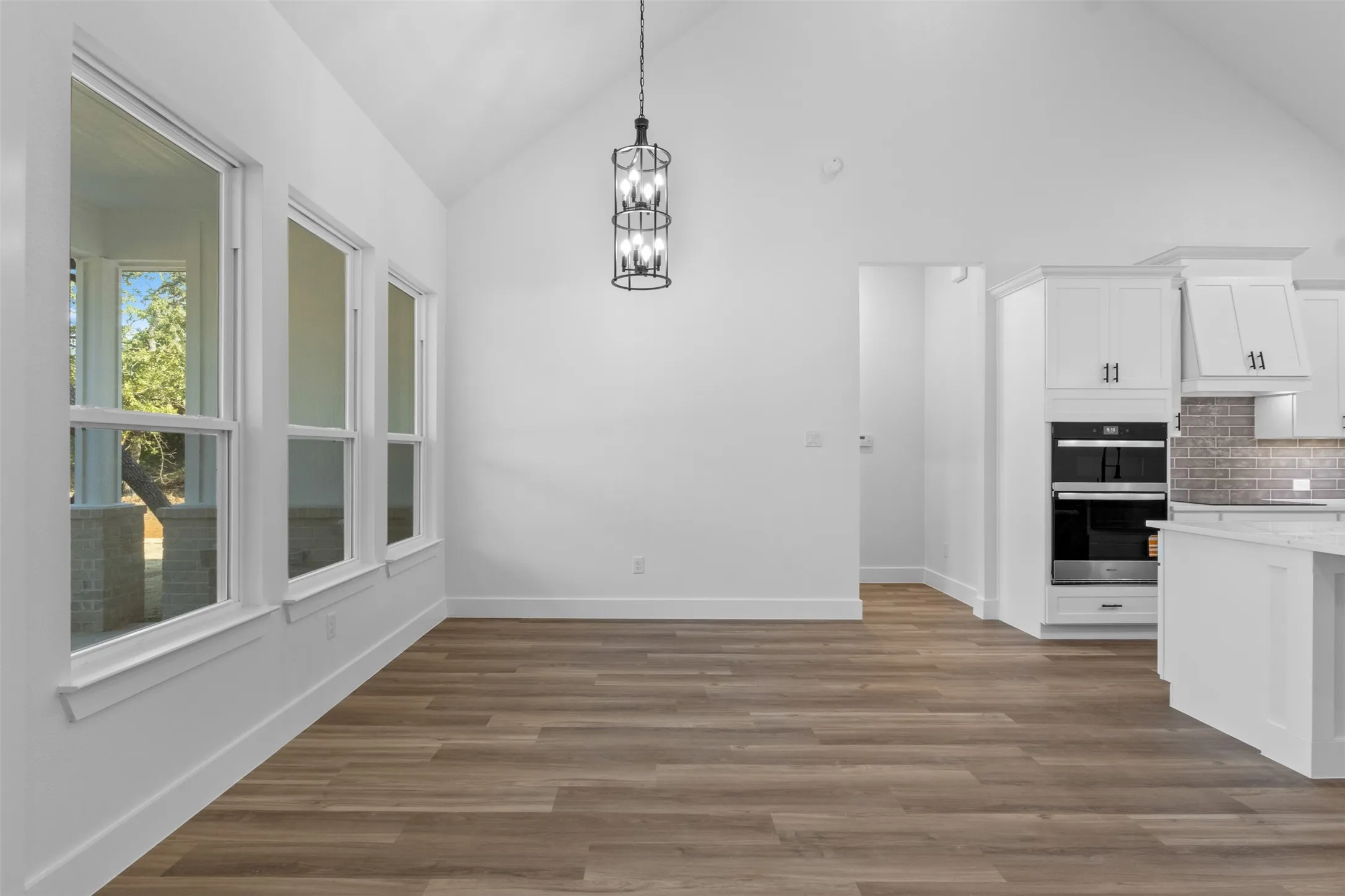 Vaulted ceiling with an architectural chandelier, wood-finish flooring, white cabinetry with dark hardware, integrated double ovens, and large windows