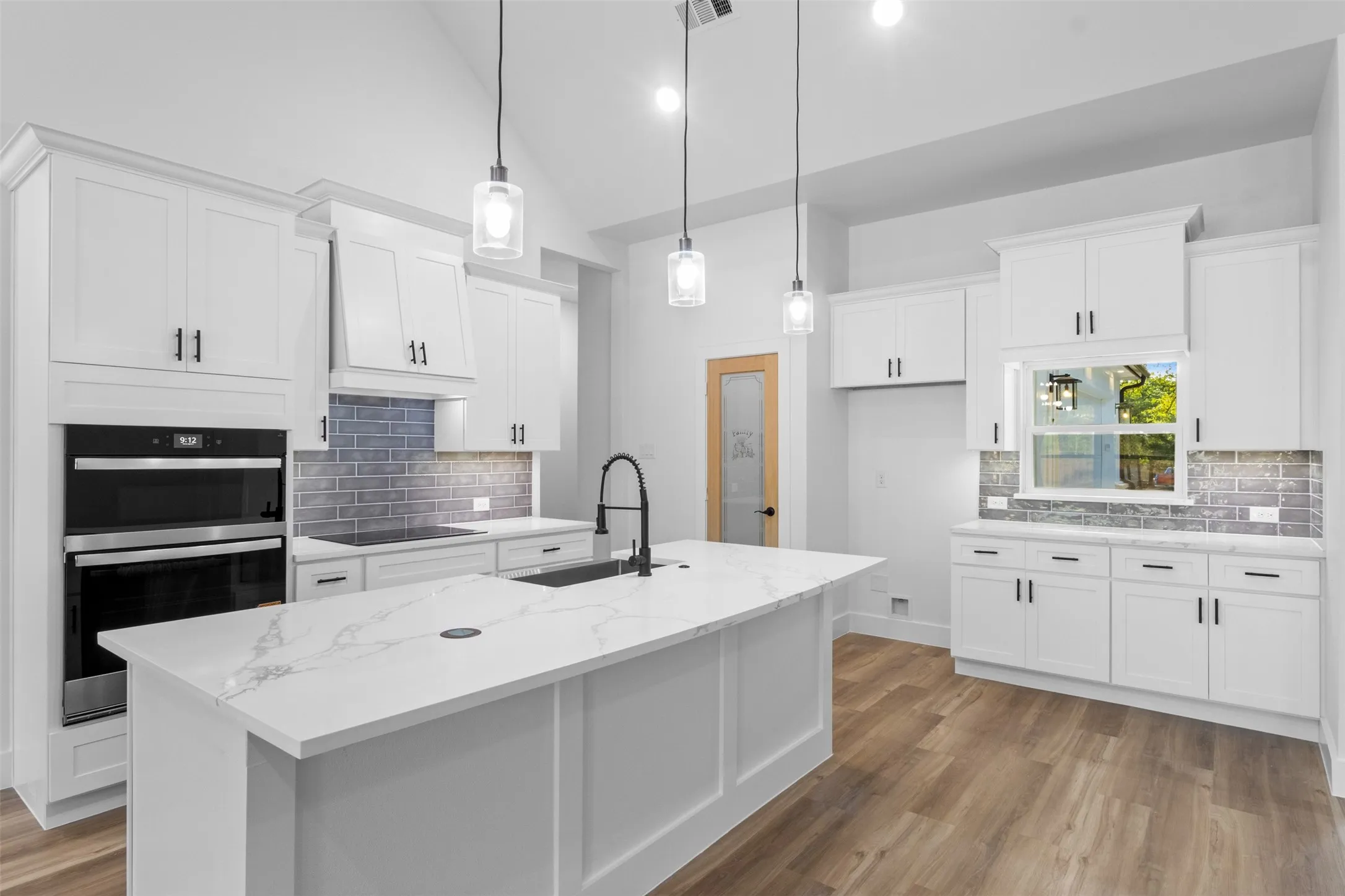 Modern kitchen featuring white shaker-style cabinetry with chrome hardware, a large waterfall-edge island with white quartz countertop, stainless steel appliances including double wall ovens, pendant lighting with glass shades, subway tile backsplash, and wood-finish flooring throughout the open layout