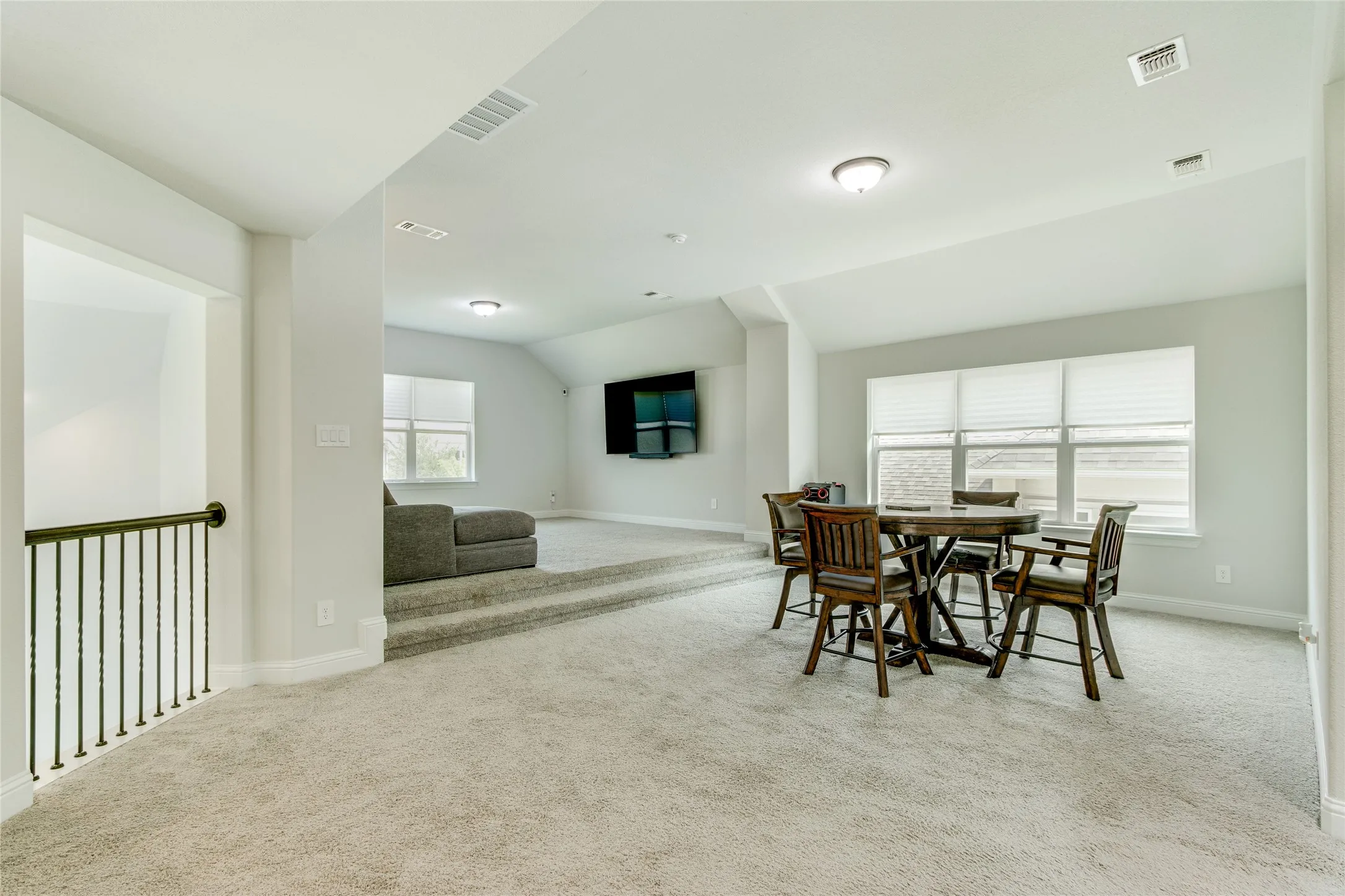Carpeted room with a stepped floor level, multiple windows with blinds, a black metal staircase railing, and recessed lighting
