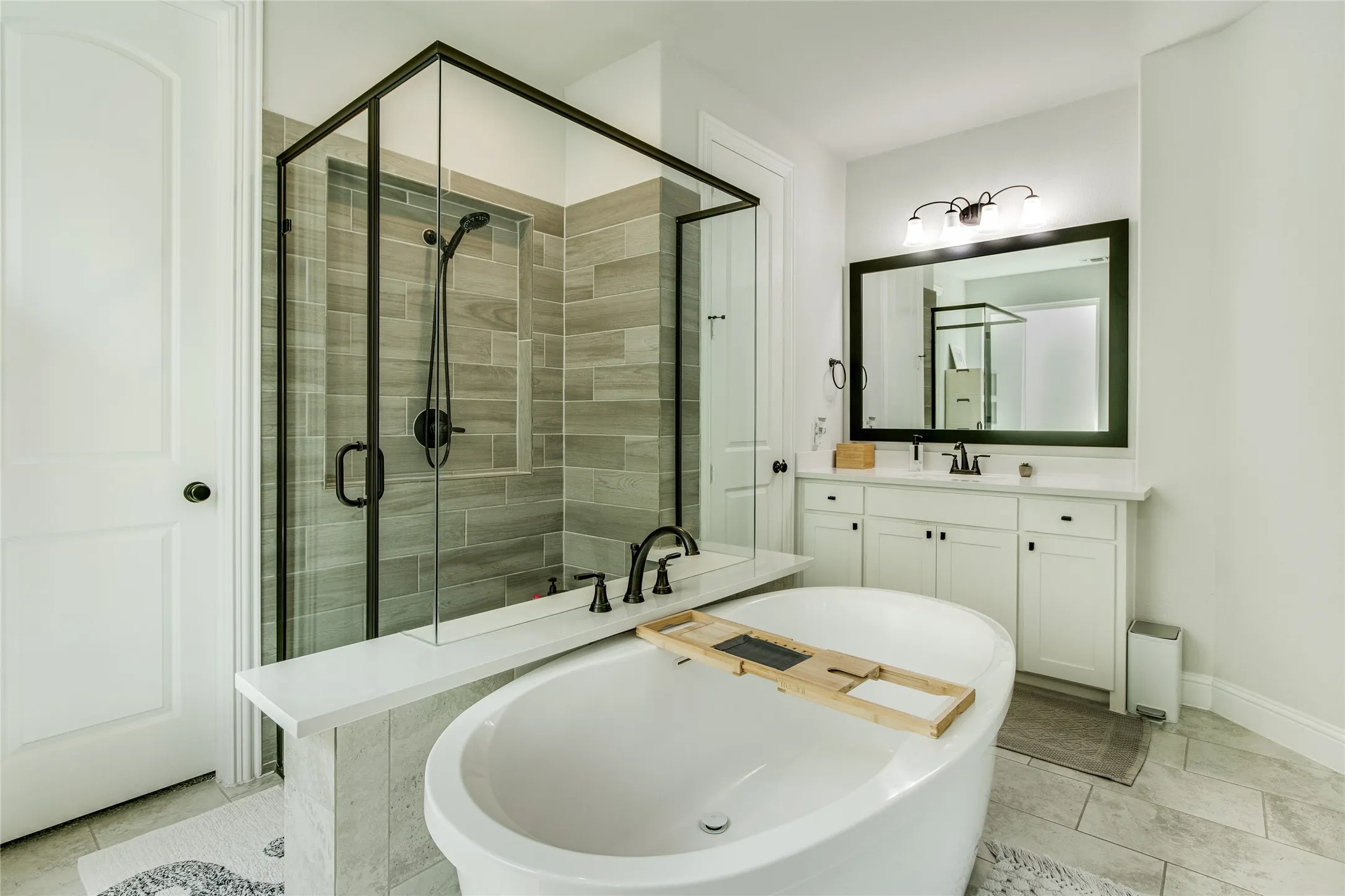 Freestanding soaking tub, glass-enclosed shower with tiled surround, dual-sink vanity with white cabinetry, large framed mirror, and tile flooring