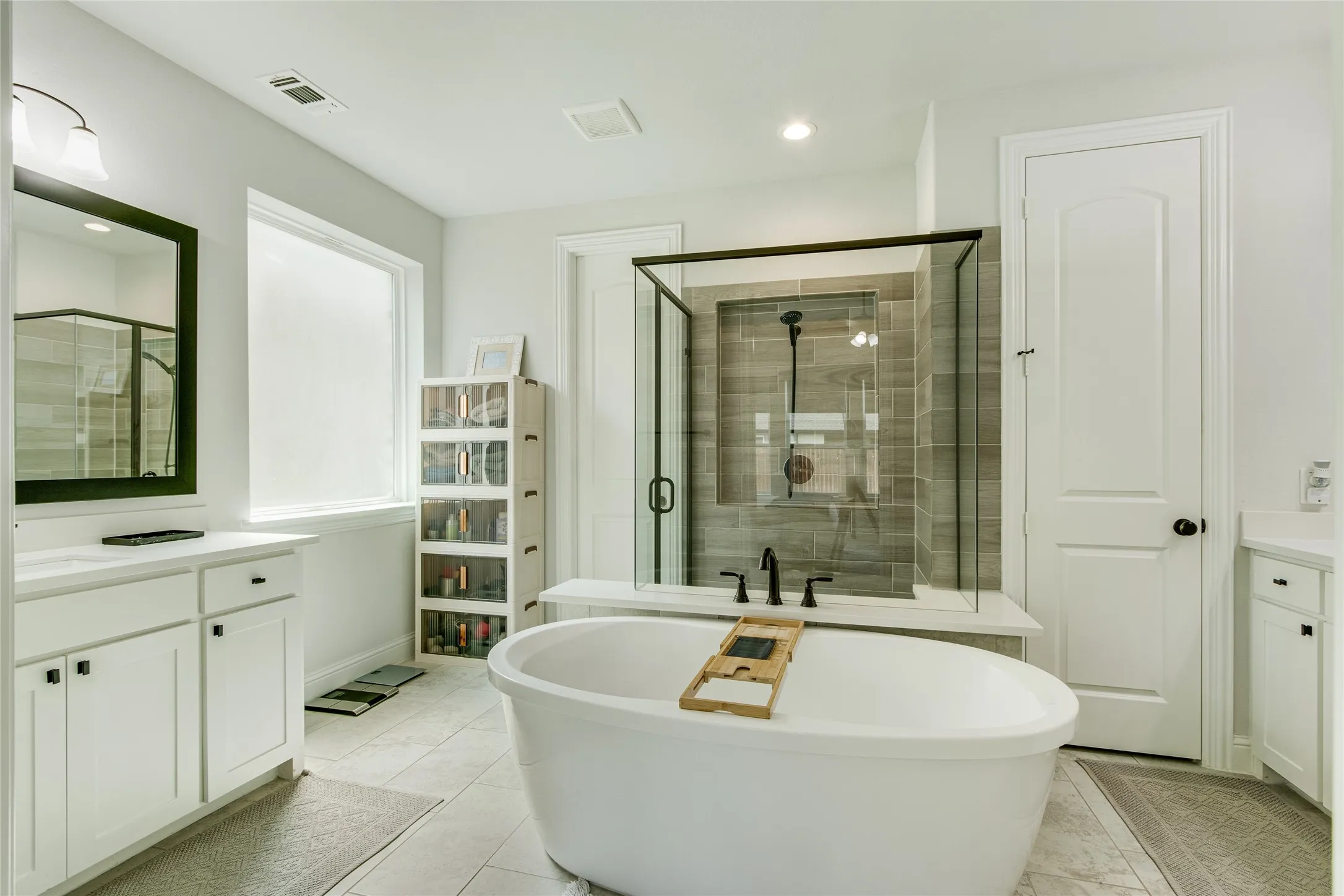 Freestanding soaking tub with black fixtures, walk-in shower with glass enclosure and tiled walls, dual vanities with white cabinetry, and large window