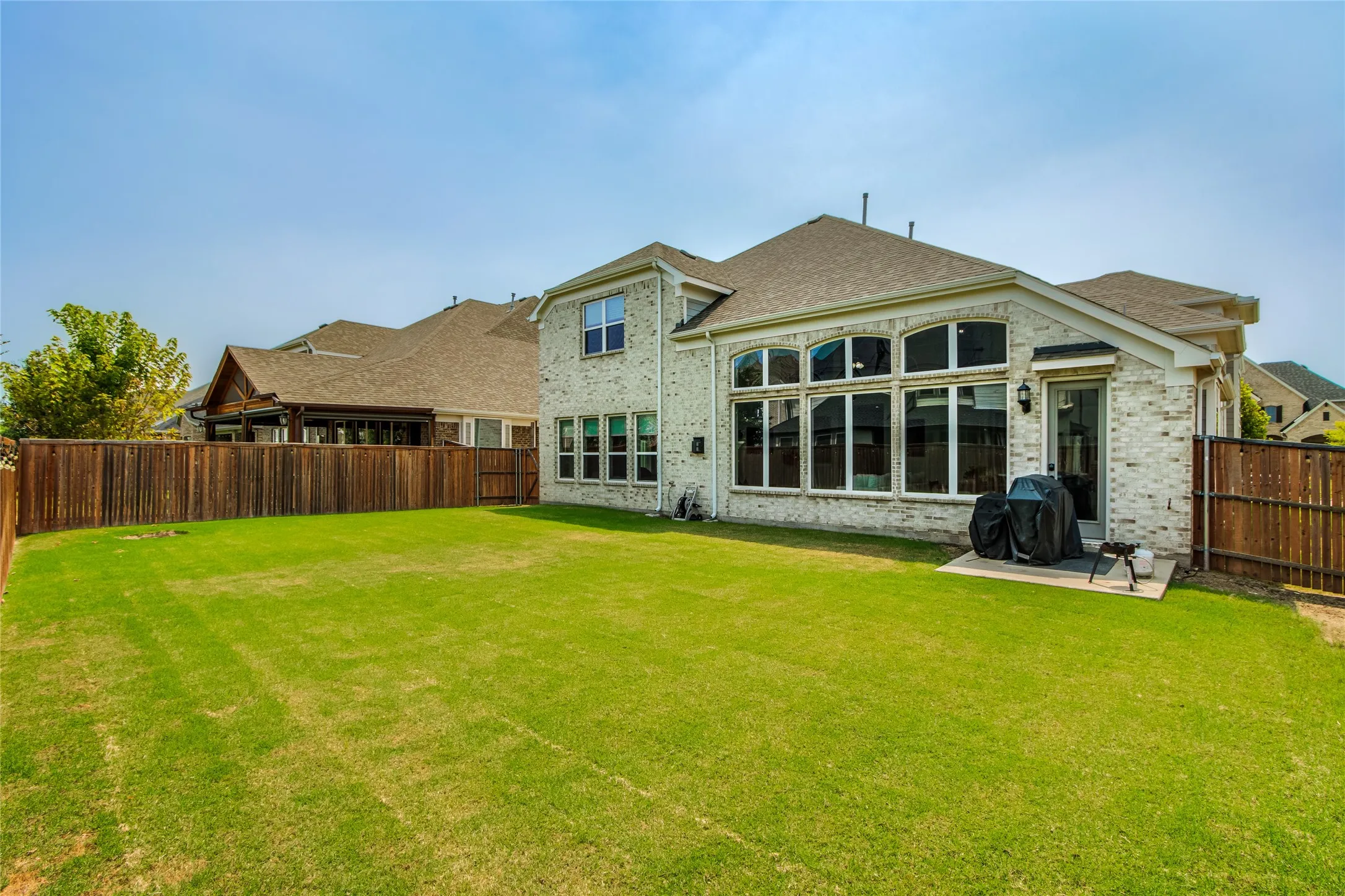 Expansive backyard featuring a manicured lawn bordered by wood-finish fencing, with a two-story brick and stone residence showcasing multiple large windows, a covered patio area, and a pitched roof with architectural detailing