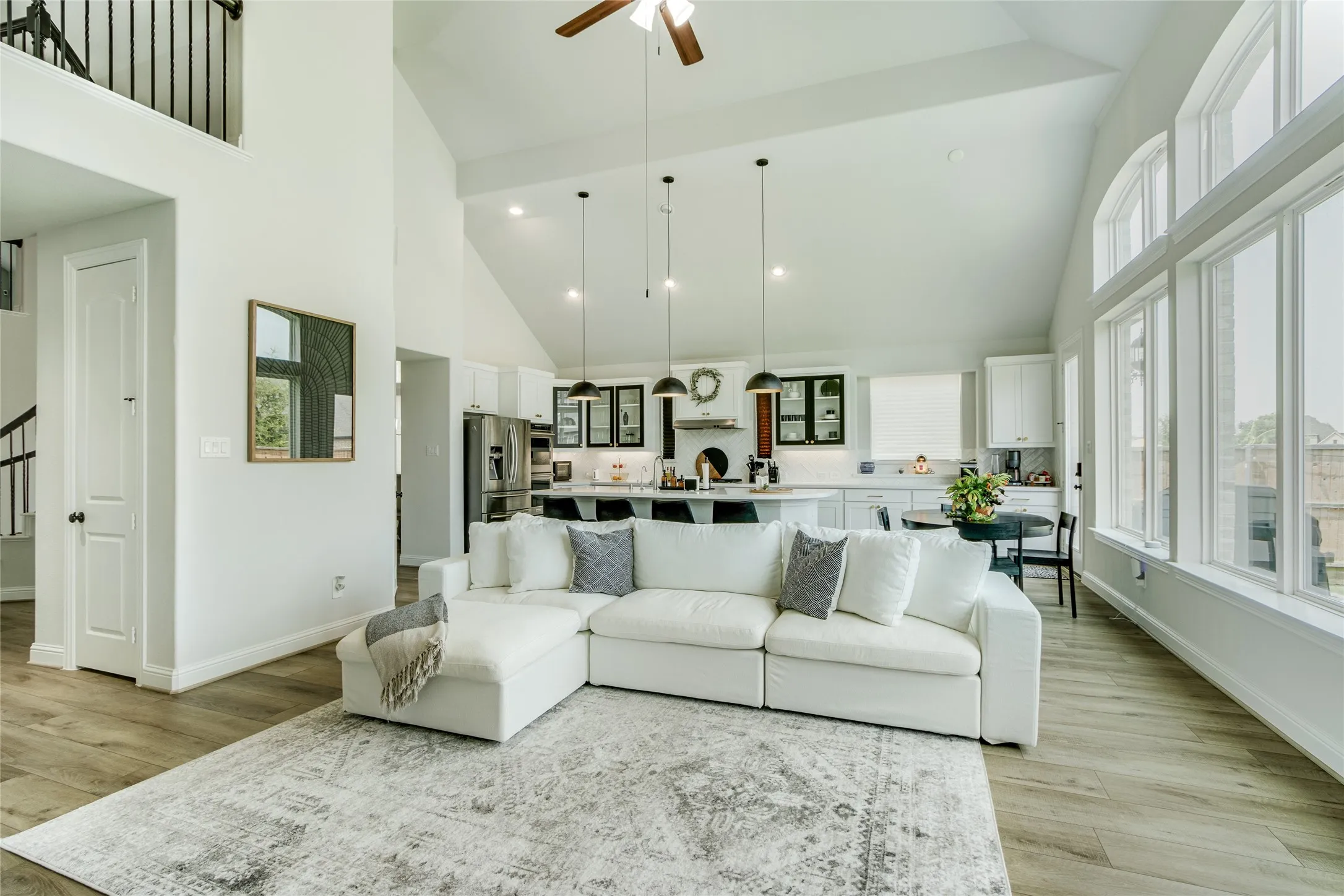 Open-concept living space featuring soaring vaulted ceilings, abundant natural light from expansive windows, hardwood flooring, a white sectional with chaise, neutral area rug, integrated kitchen with white cabinetry and stainless steel appliances, pendant lighting, and a ceiling fan